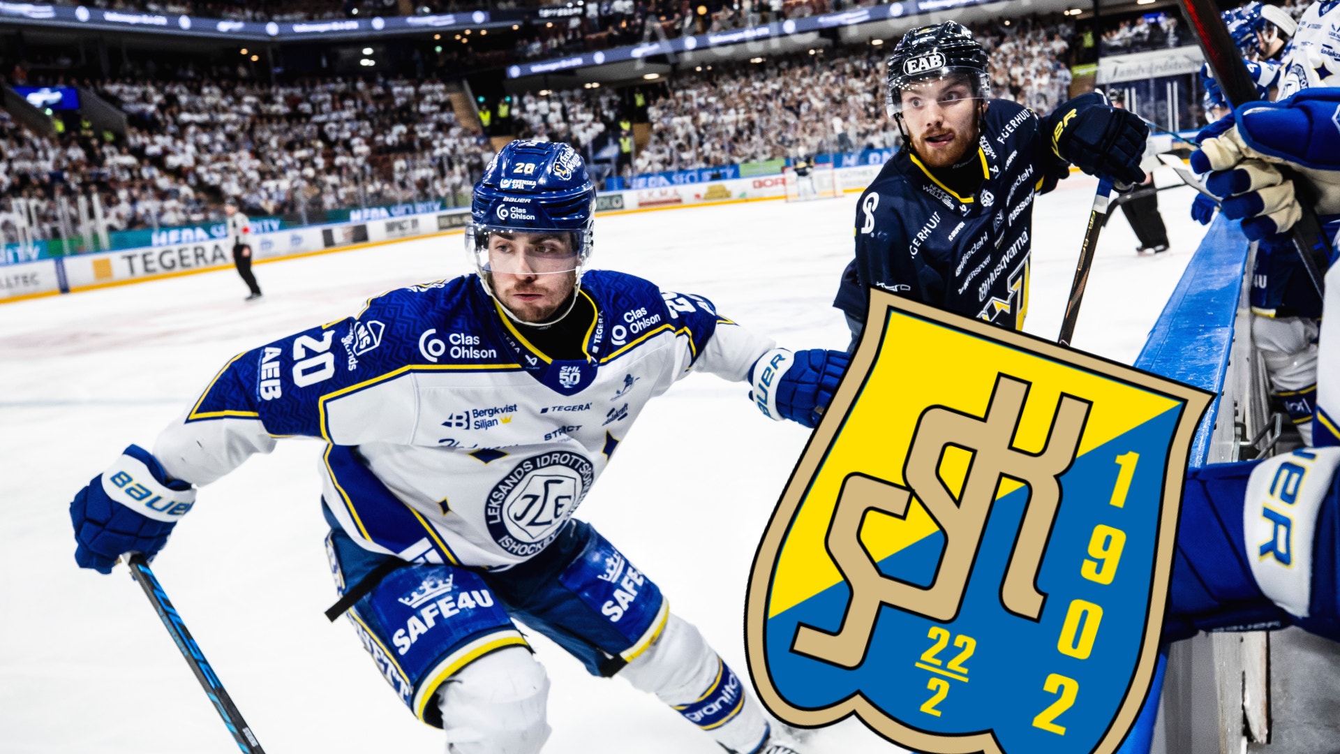 Ishockeymatch med spelare i blå och vit tröja som skär över isen nära banden med publikhall i bakgrunden.