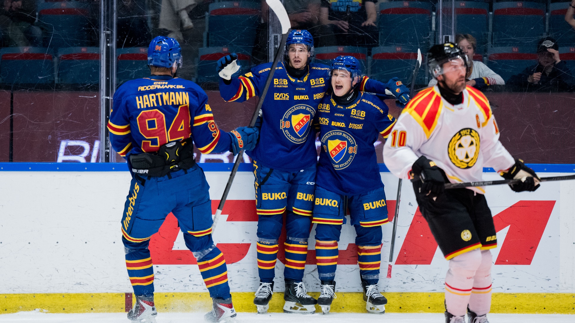 Ishockeymatch med spelare i blåa och vita trikåer på isen. Flera spelare står tillsammans på isen under en match, med publik synlig i bakgrunden.
