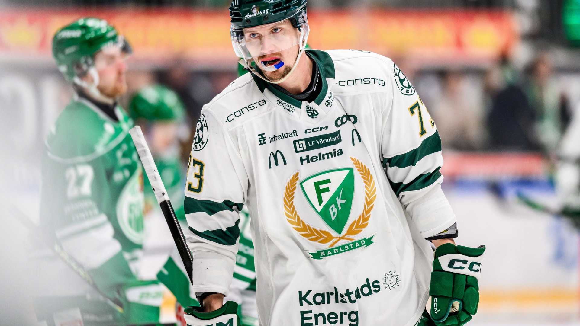 Ishockeylaget sitter på spelarnas bänk under en match. Spelarna bär gula tröjor med svart och röd detaljer, svarta hjälmar och vita handskar. De sitter tätt tillsammans och följer matchen.