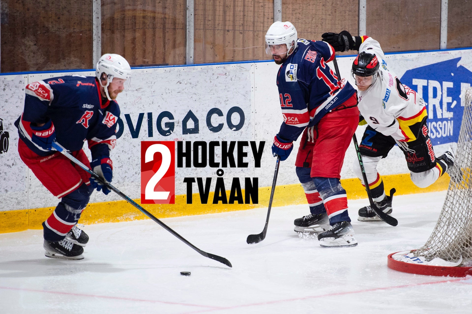 Ishockeymatch med spelare från två lag i aktion på isen. Spelare i mörkblå och röda trikåer spelar mot ett lag i vitt och gult. En spelare håller i sin hockeyklubb nära pucken medan lagkamrater positionerar sig runt målet.
