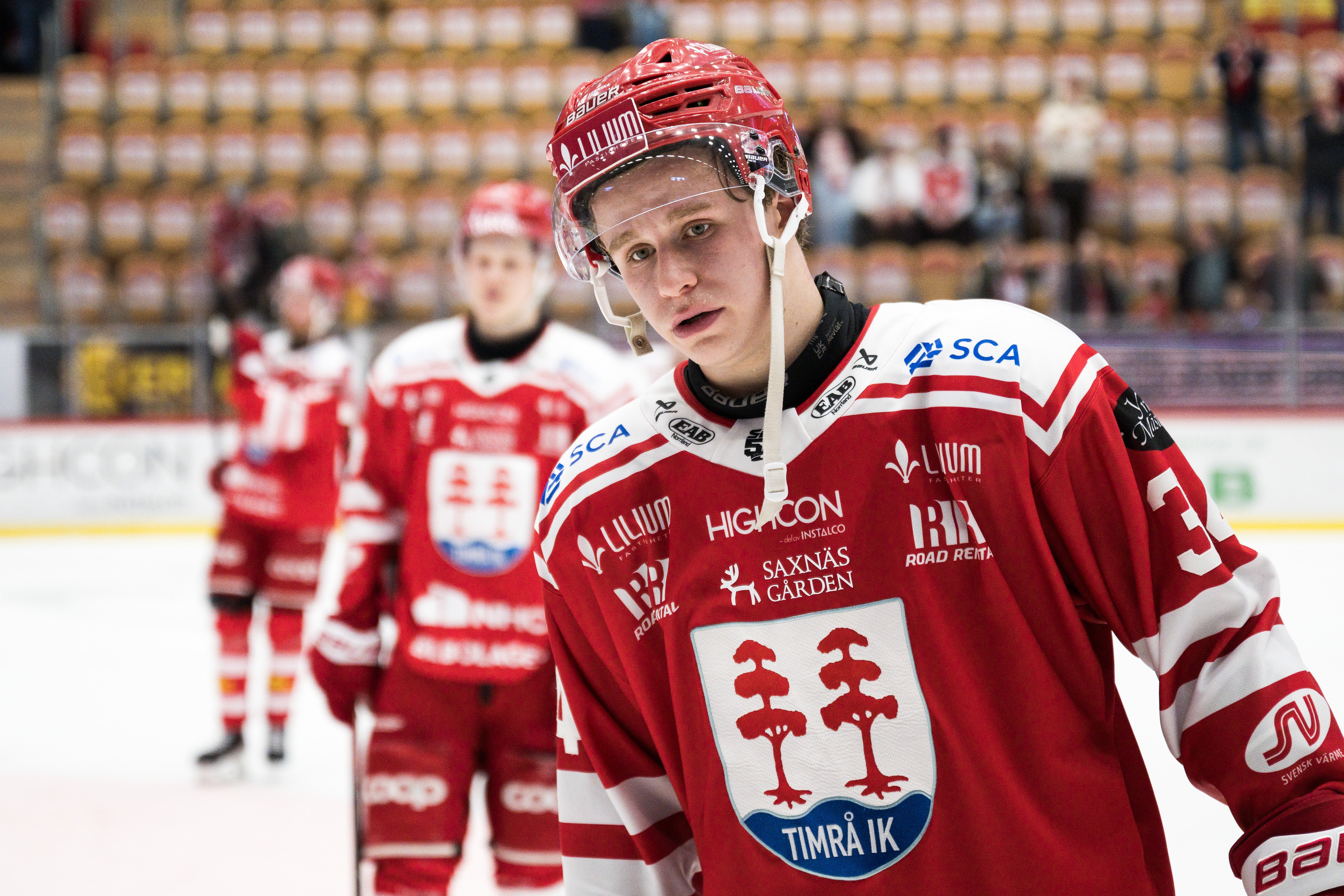 Ishockeyspelare i röd och vit matchdräkt med hjälm på en ishockeyplan med publik i bakgrunden.