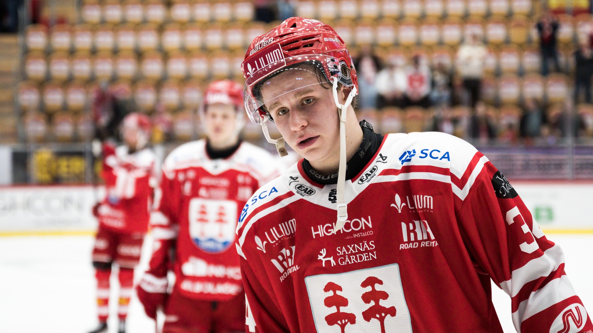 Ishockeyspelare i röd och vit matchdräkt med hjälm på en ishockeyplan med publik i bakgrunden.