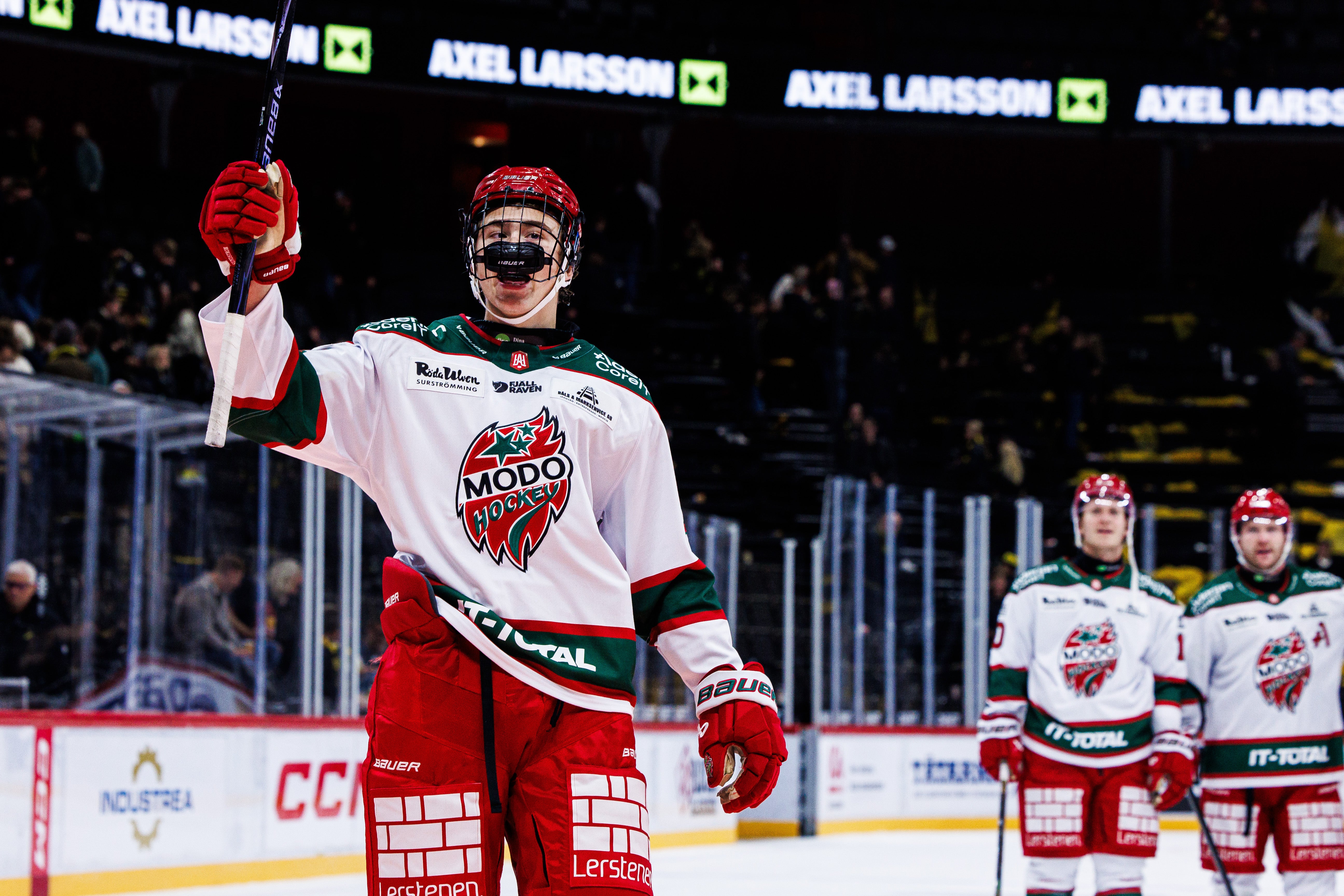 En ishockeyspelare i MODO Hockeys vit-röda matchtröja håller upp ena armen och klubban i luften på isen. Två lagkamrater står i bakgrunden i arenan.