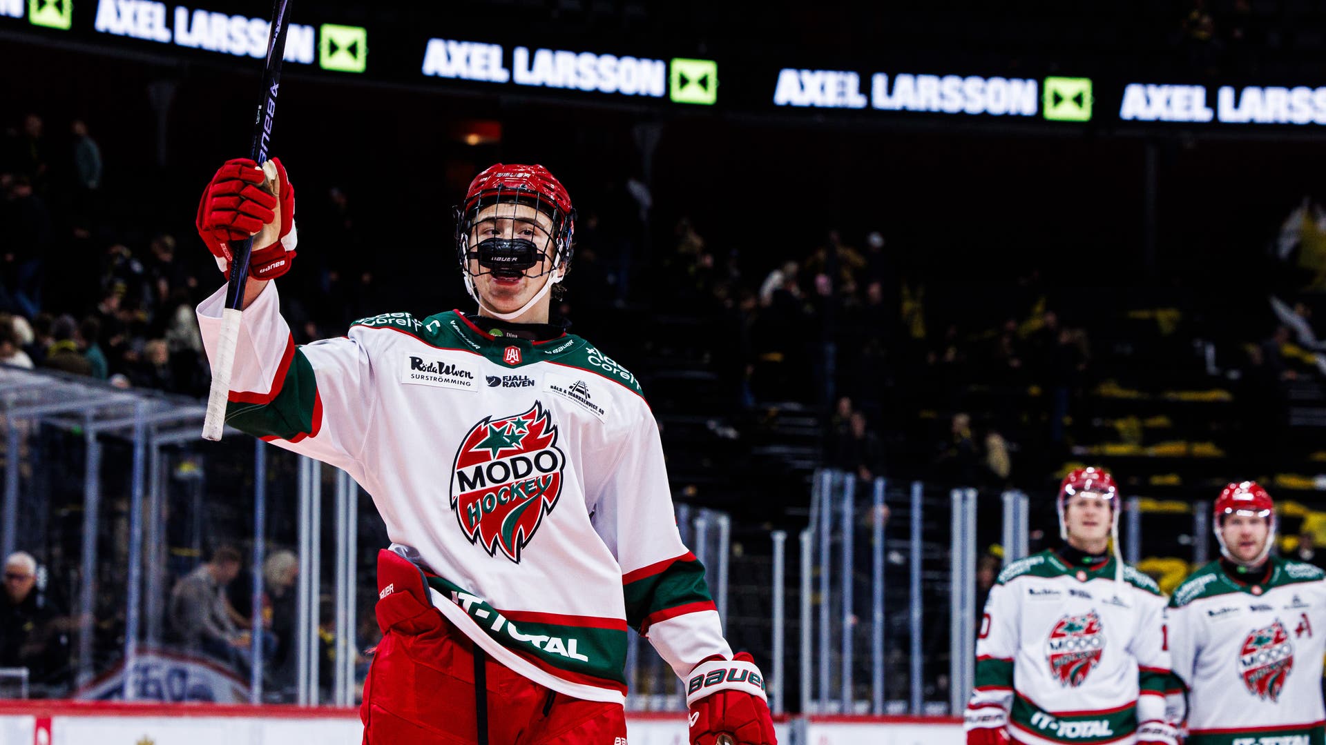 En ishockeyspelare i MODO Hockeys vit-röda matchtröja håller upp ena armen och klubban i luften på isen. Två lagkamrater står i bakgrunden i arenan.