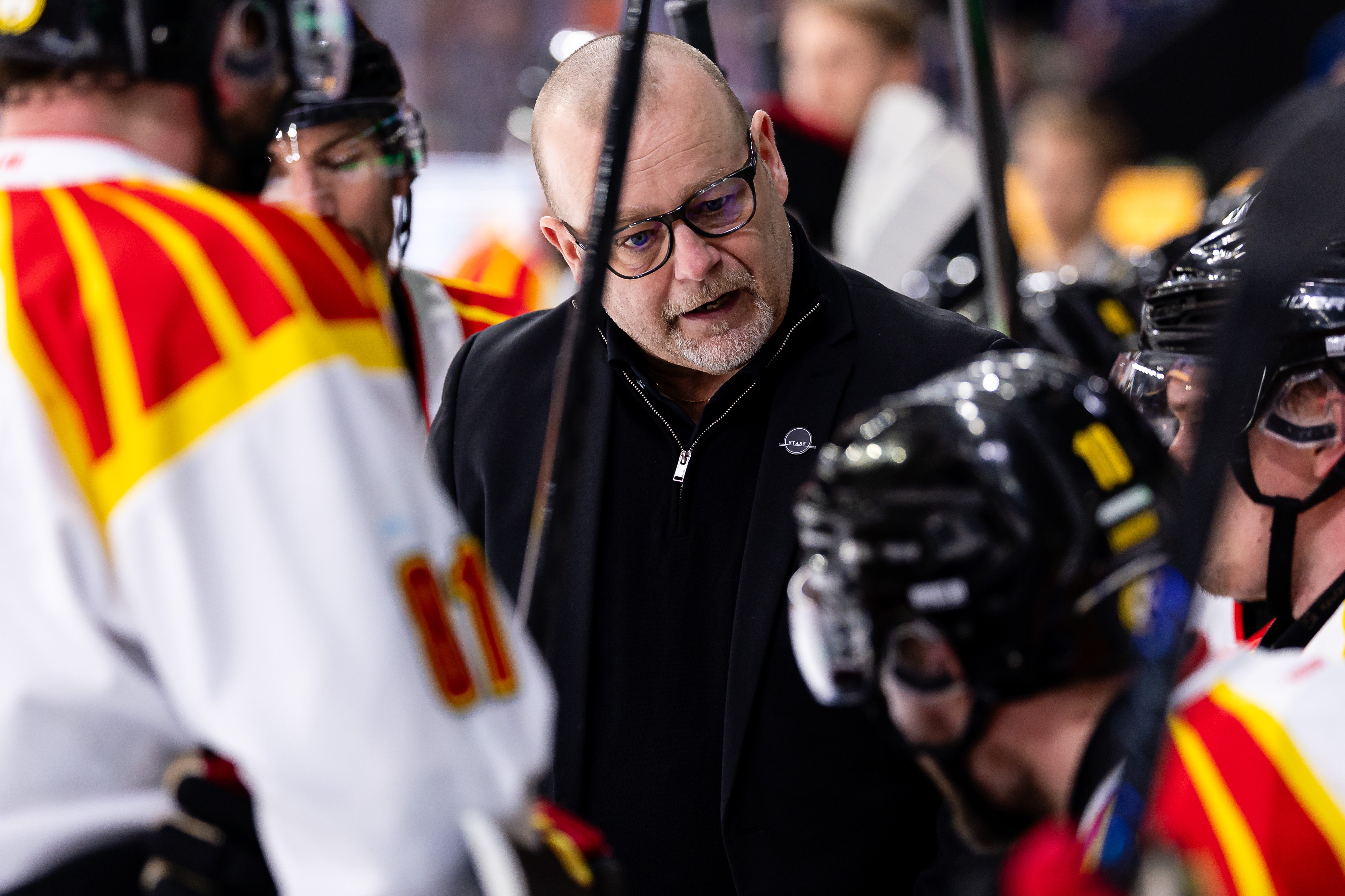 En man med glasögon i svart jacka på ishockeyns bänk omgiven av spelare i röd och gul utrustning.