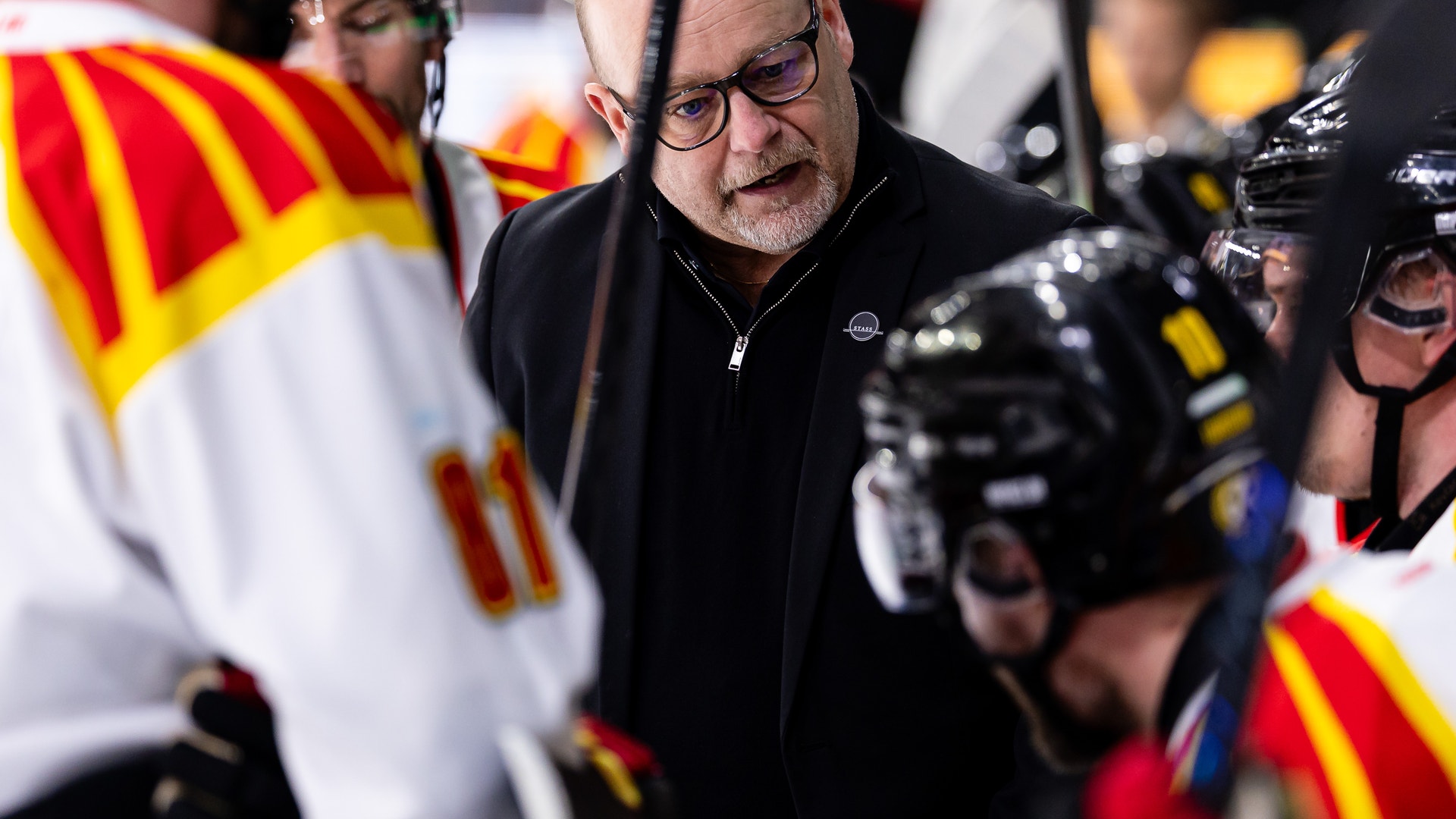 En man med glasögon i svart jacka på ishockeyns bänk omgiven av spelare i röd och gul utrustning.