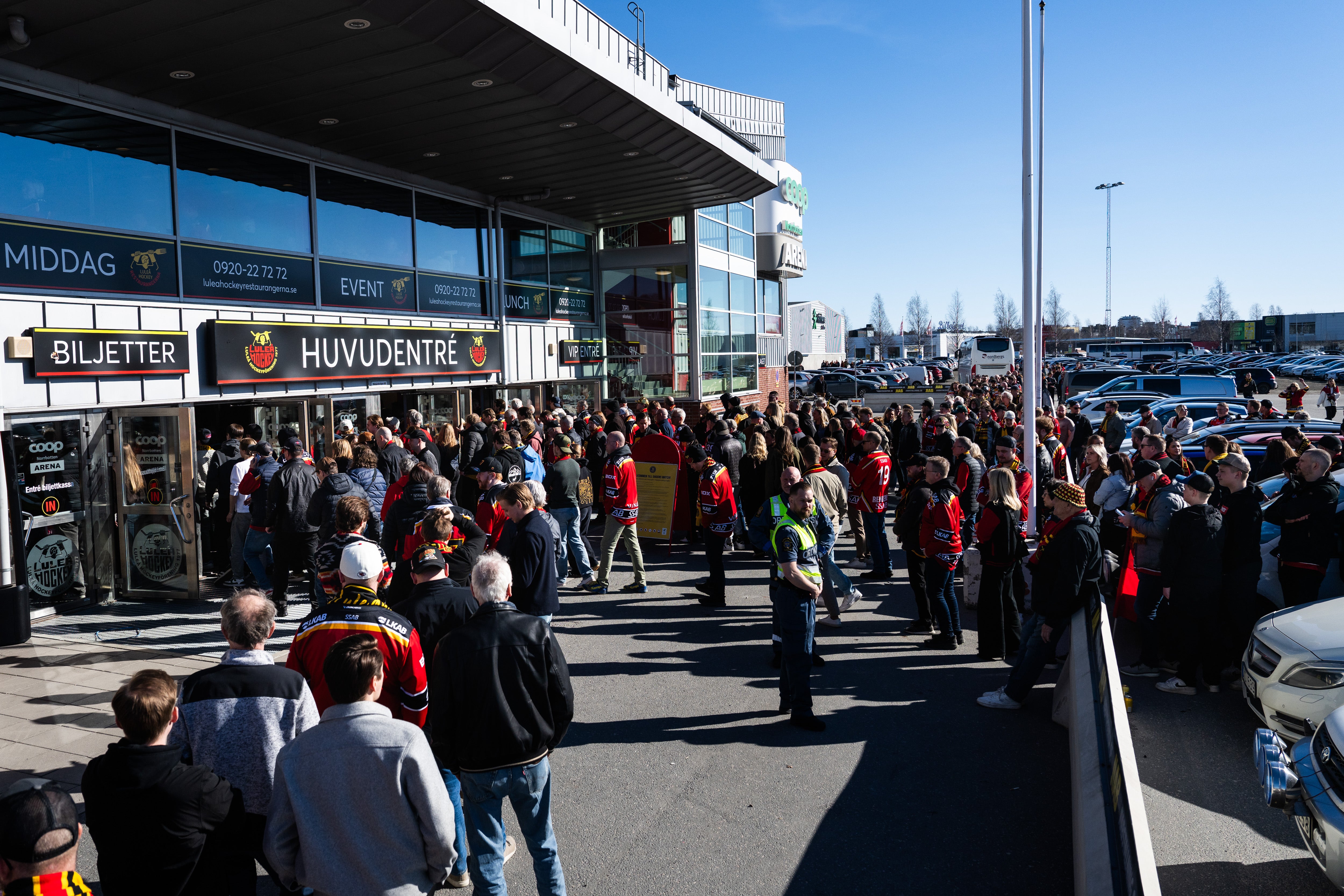 En stor folksamling står utanför en ishall eller sportanläggning på en solig dag. Människor väntar vid huvudentrén och biljettluckan.