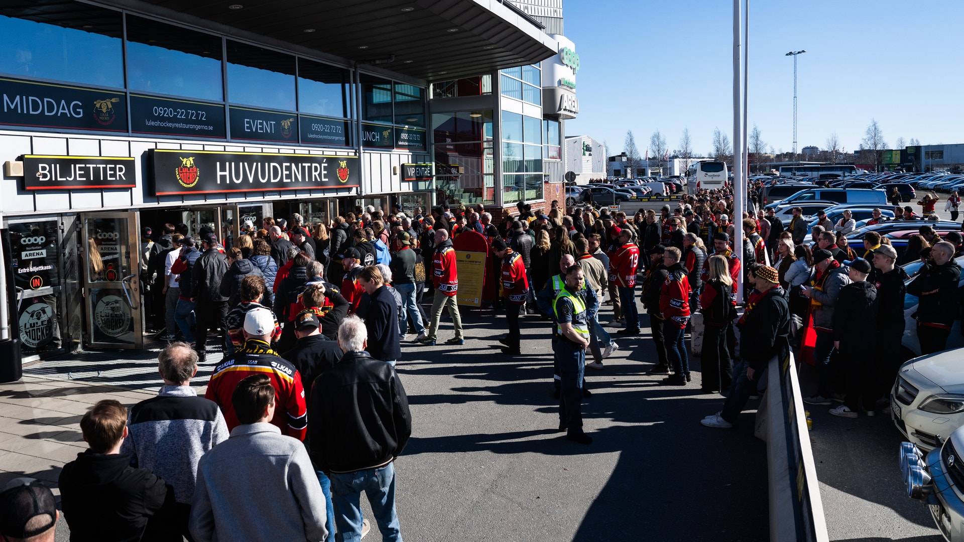 En stor folksamling står utanför en ishall eller sportanläggning på en solig dag. Människor väntar vid huvudentrén och biljettluckan.