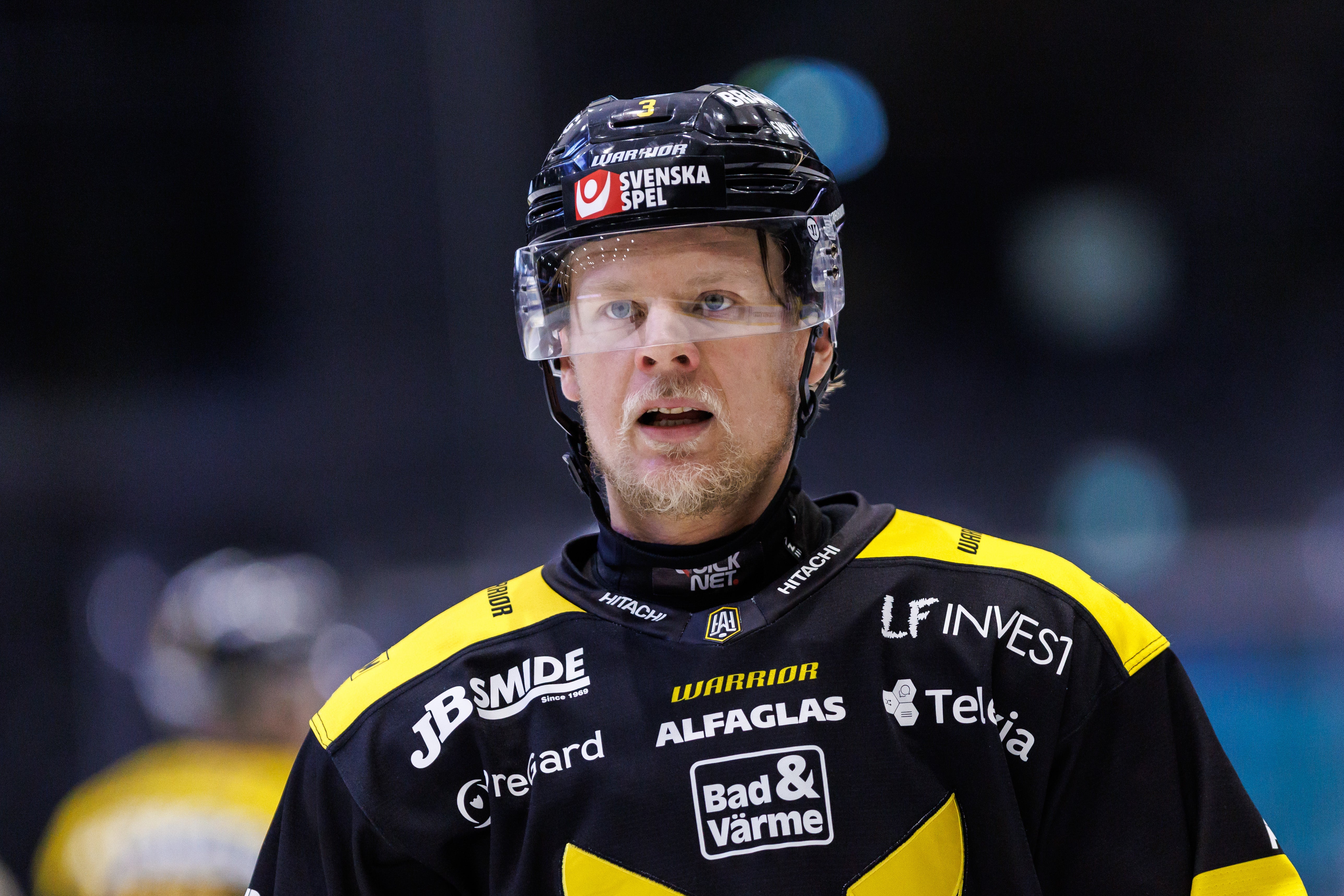Ishockeyspelare i svart och gul tröja med sponsorlogotyper, svart hjälm och skyddsglasögon, fotograferad under match.