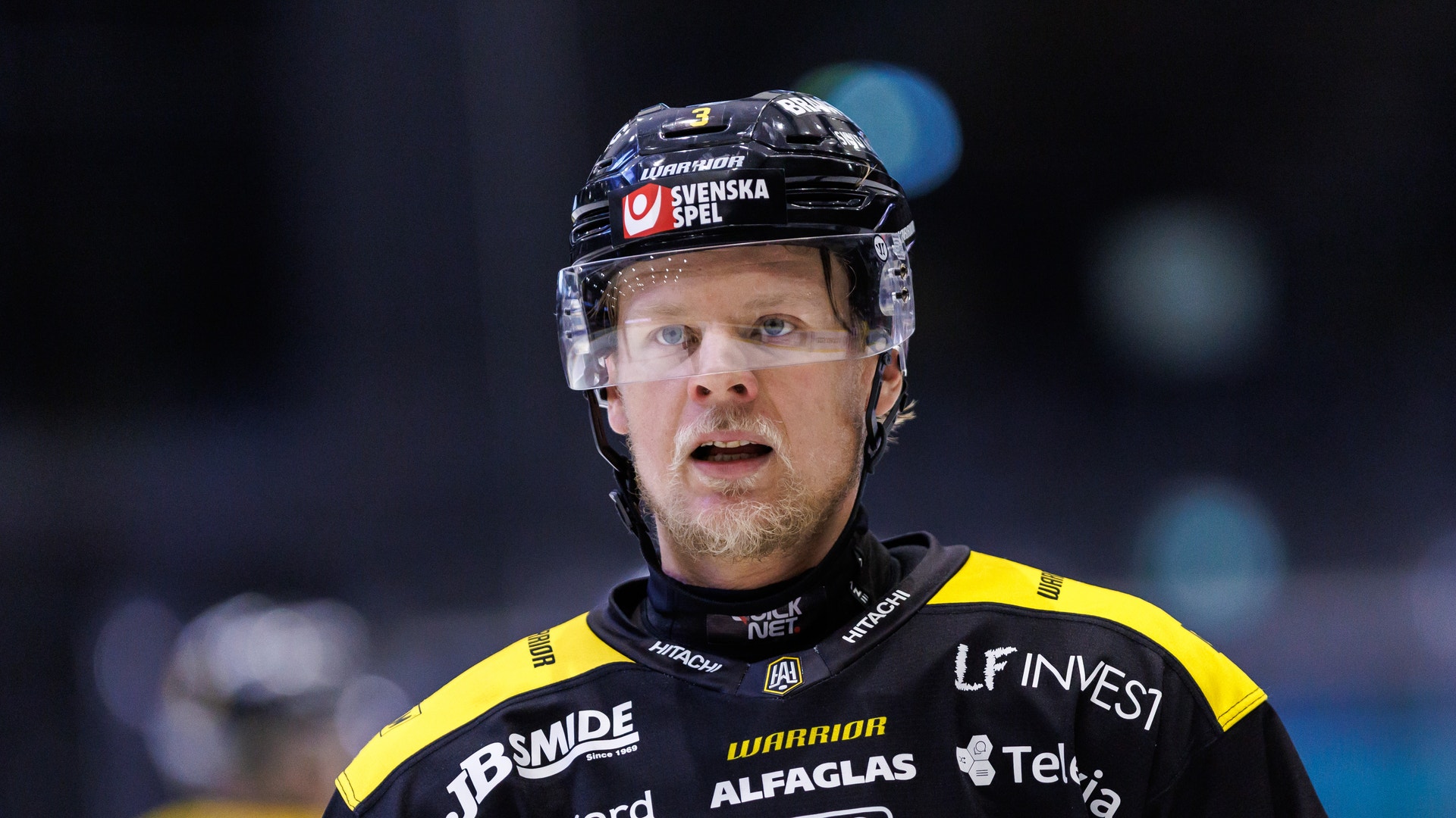Ishockeyspelare i svart och gul tröja med sponsorlogotyper, svart hjälm och skyddsglasögon, fotograferad under match.