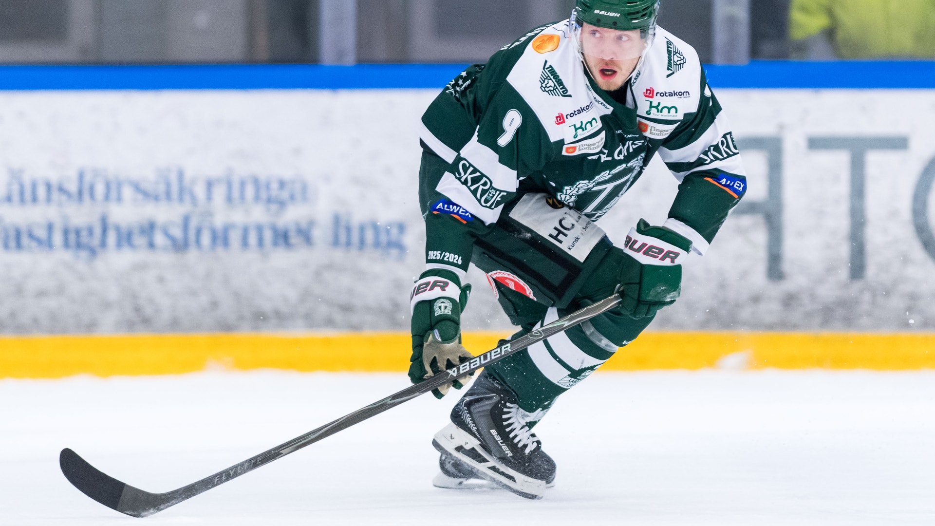 Ishockeyspelare i grön och vit tröja spelar på ishockeyrink med hockeyklubbans sponsorlogotyper på tröjan. Spelaren är i rörelse med hockeyklubbor och är fokuserad på spelet.