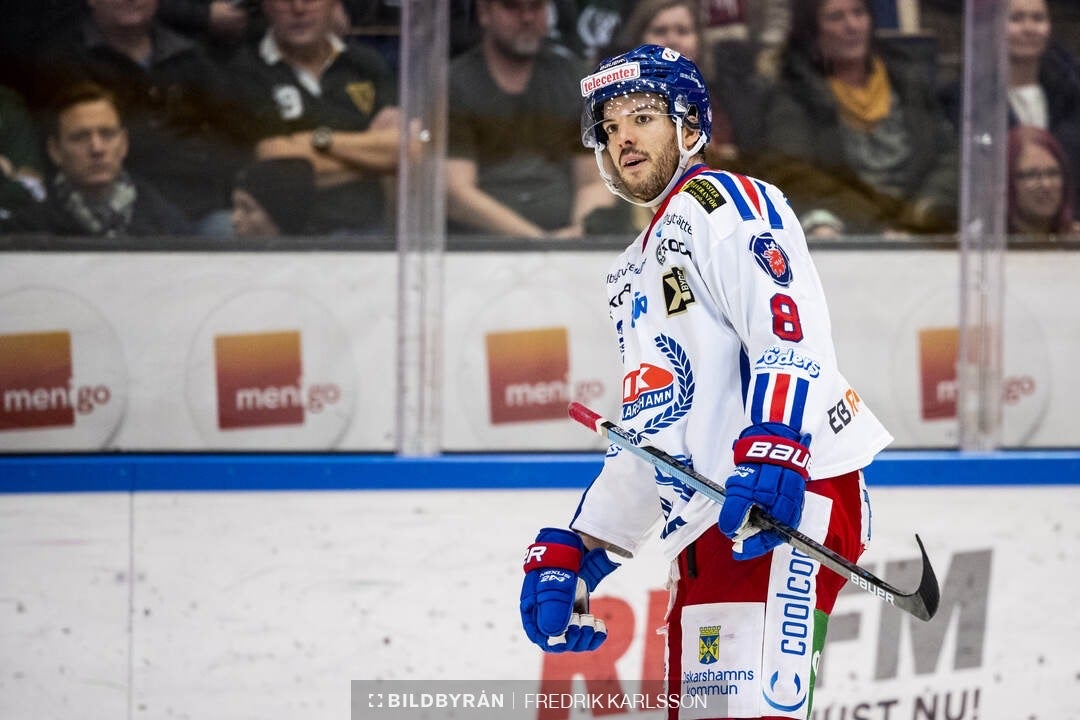 Ishockeyspelare i vit tröja med nummer 8 under en match, skådar mot kameran med publiken i bakgrunden.