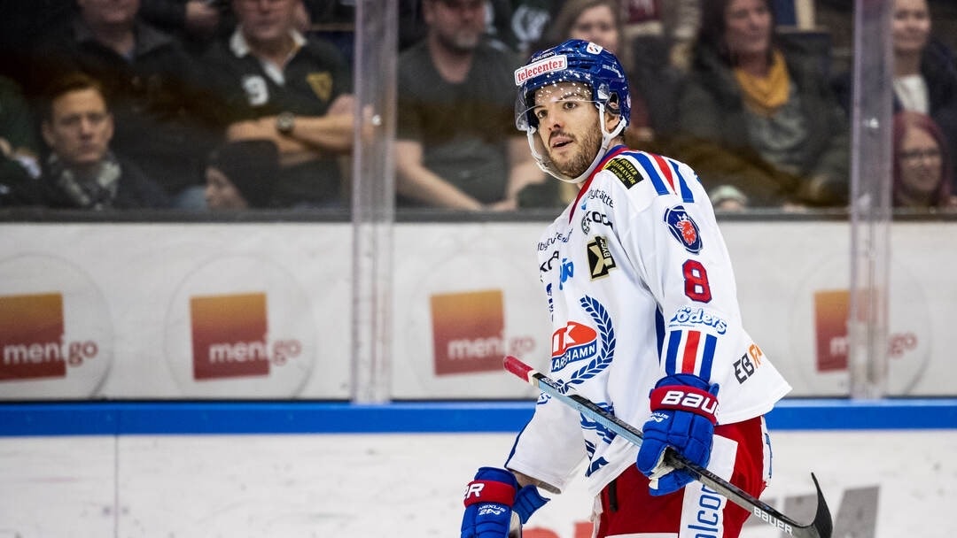 Ishockeyspelare i vit tröja med nummer 8 under en match, skådar mot kameran med publiken i bakgrunden.