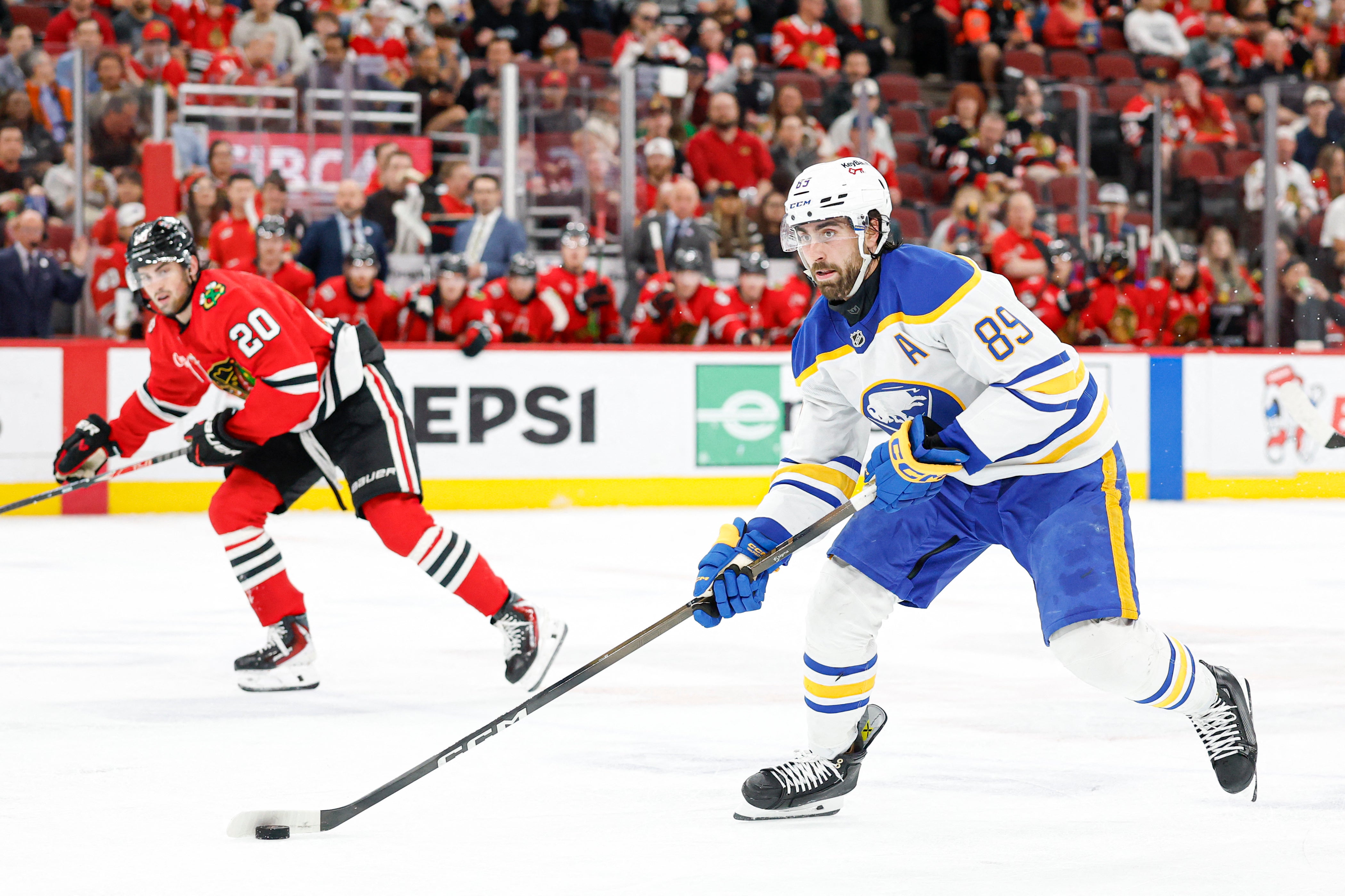 Två ishockeyspelare under en match. Spelaren i blå tröja med nummer 85 kontrollerar pucken med sin stick, medan en spelare i röd tröja med nummer 20 försöker försvara. Publiken syns i bakgrunden.