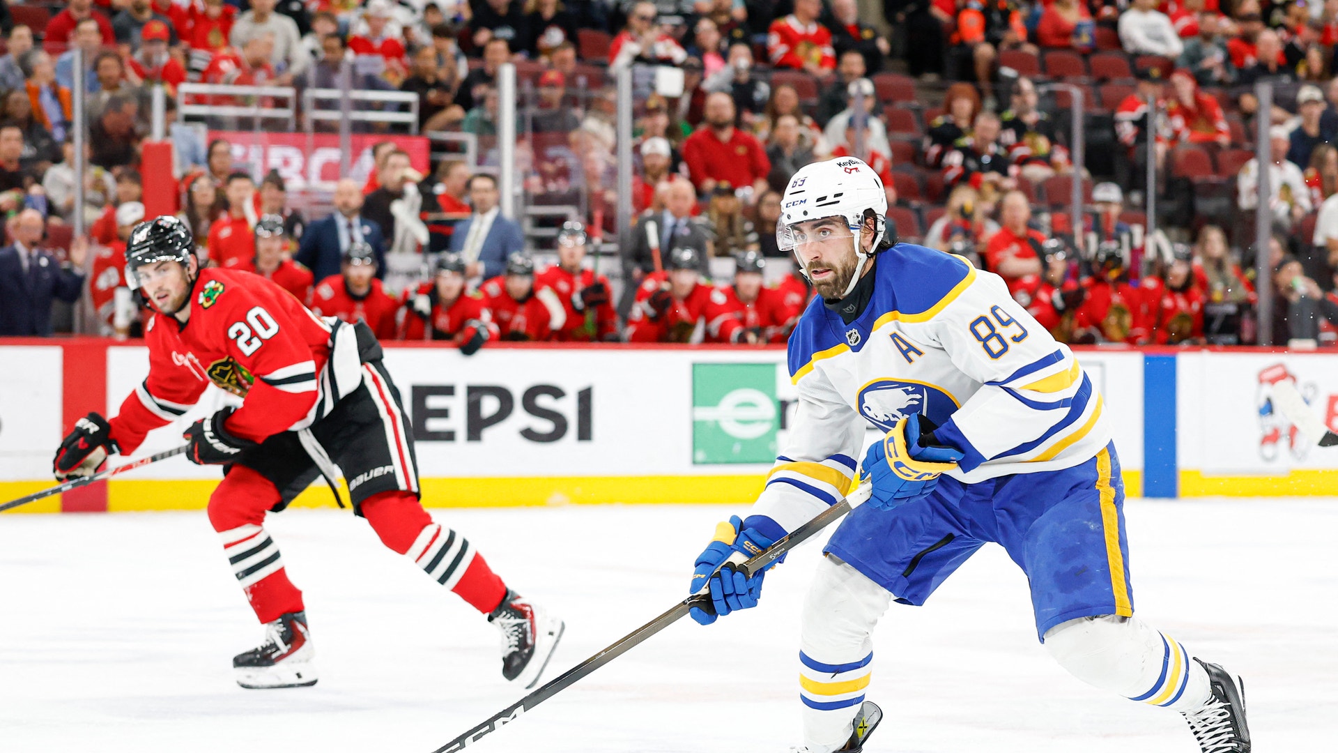 Två ishockeyspelare under en match. Spelaren i blå tröja med nummer 85 kontrollerar pucken med sin stick, medan en spelare i röd tröja med nummer 20 försöker försvara. Publiken syns i bakgrunden.