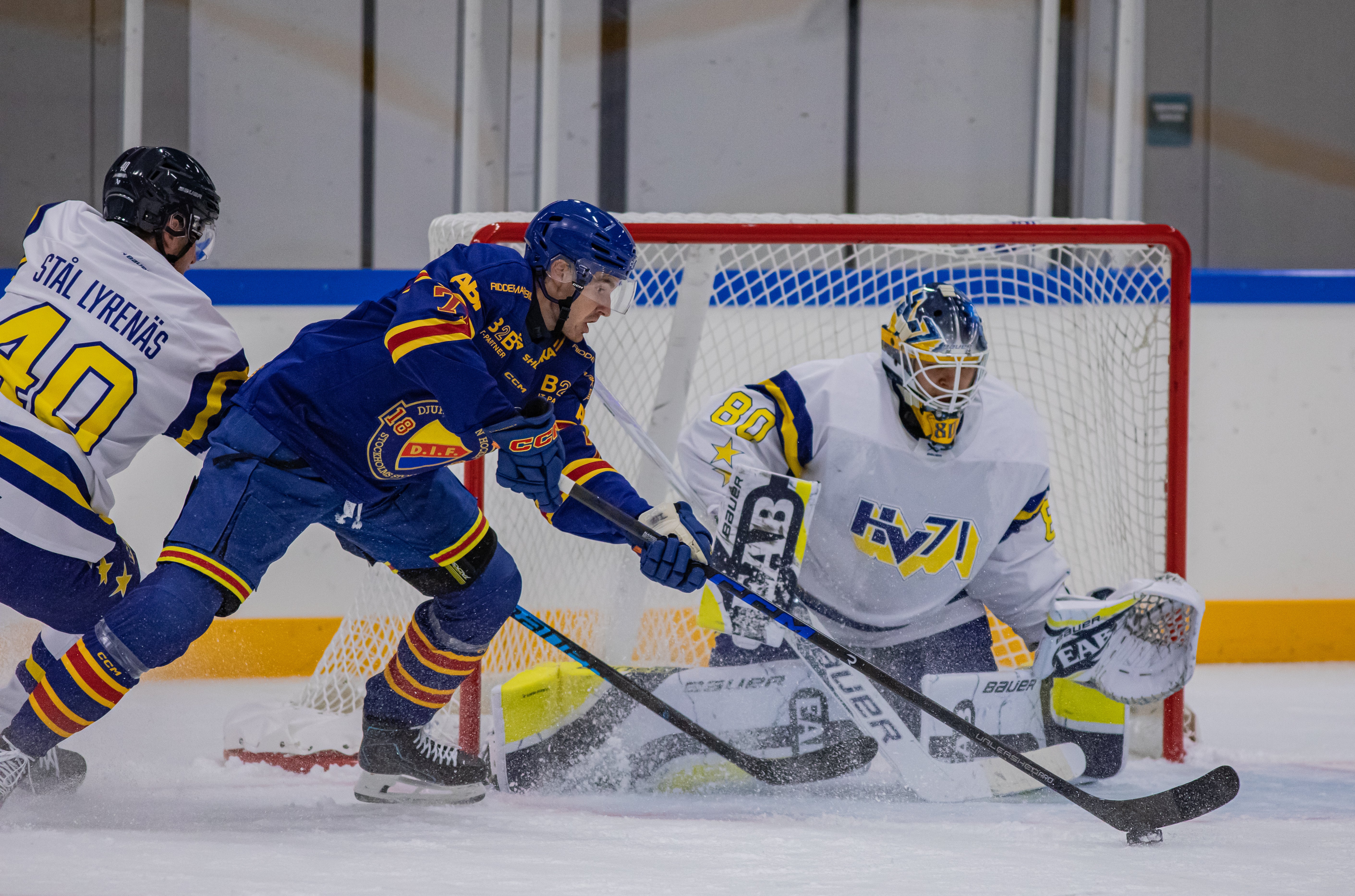 Ishockeymatch där en målvakt i vit utrustning försvarar målet mot en anfallande spelare i blå och orange tröja.