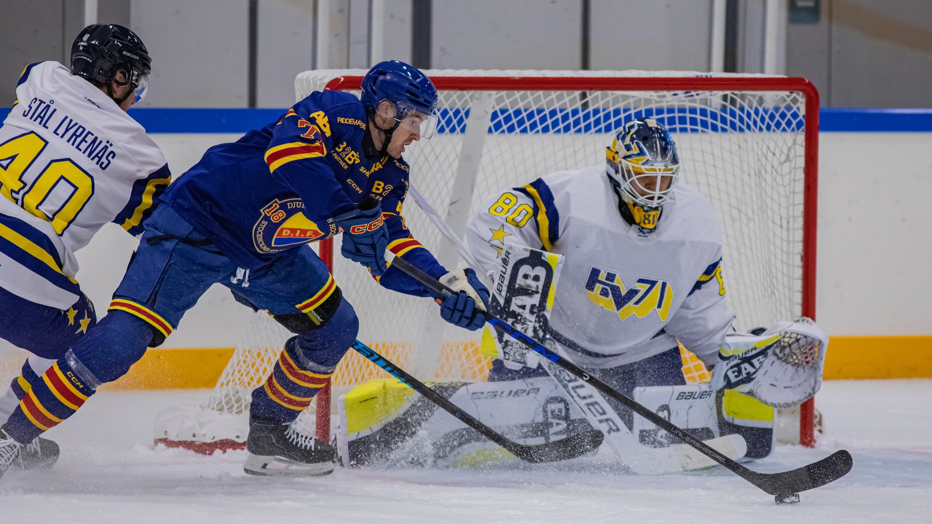 Ishockeymatch där en målvakt i vit utrustning försvarar målet mot en anfallande spelare i blå och orange tröja.