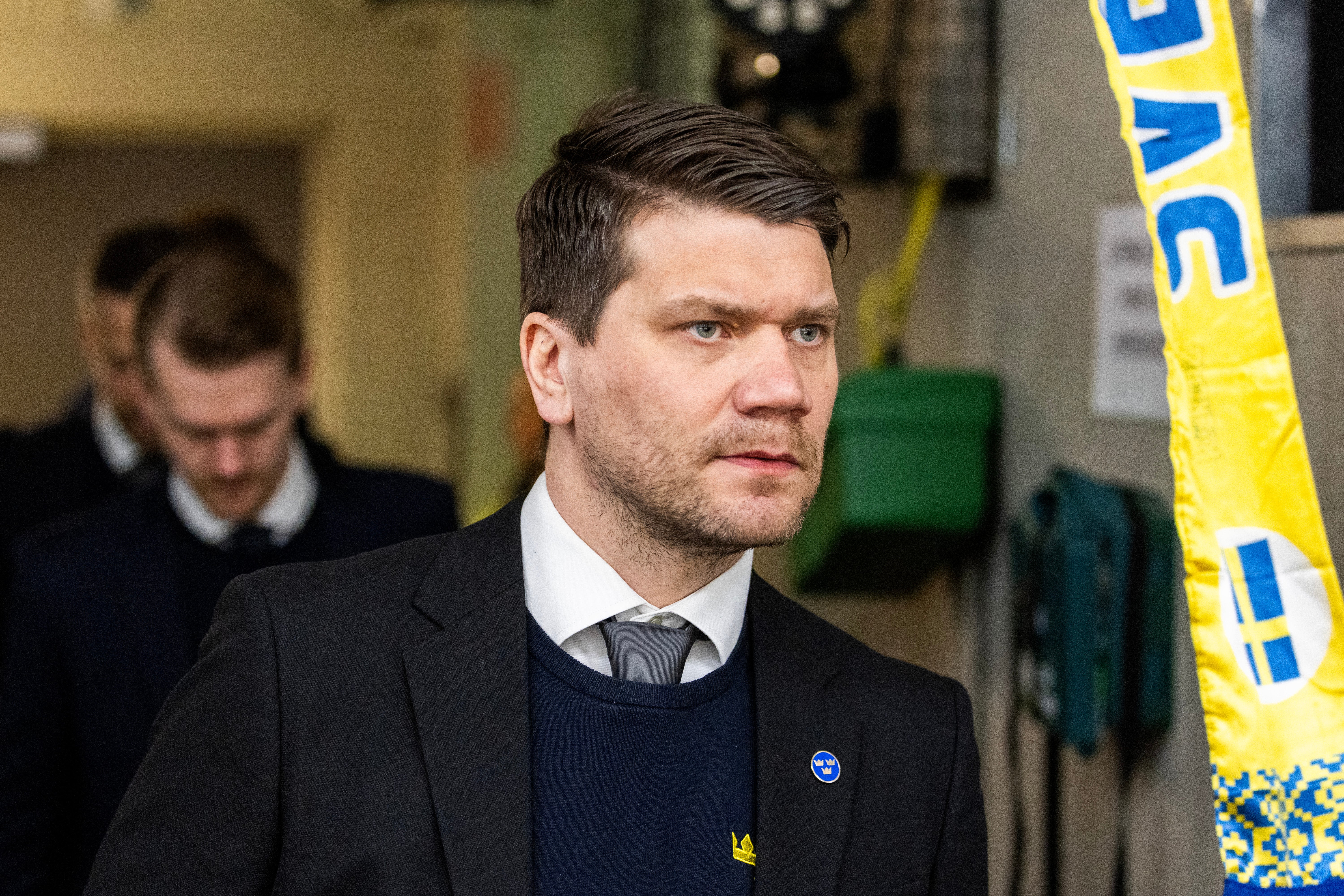 Man i mörk kostym och blå tröja står inomhus framför en gul och blå banderoll. I bakgrunden syns andra personer.