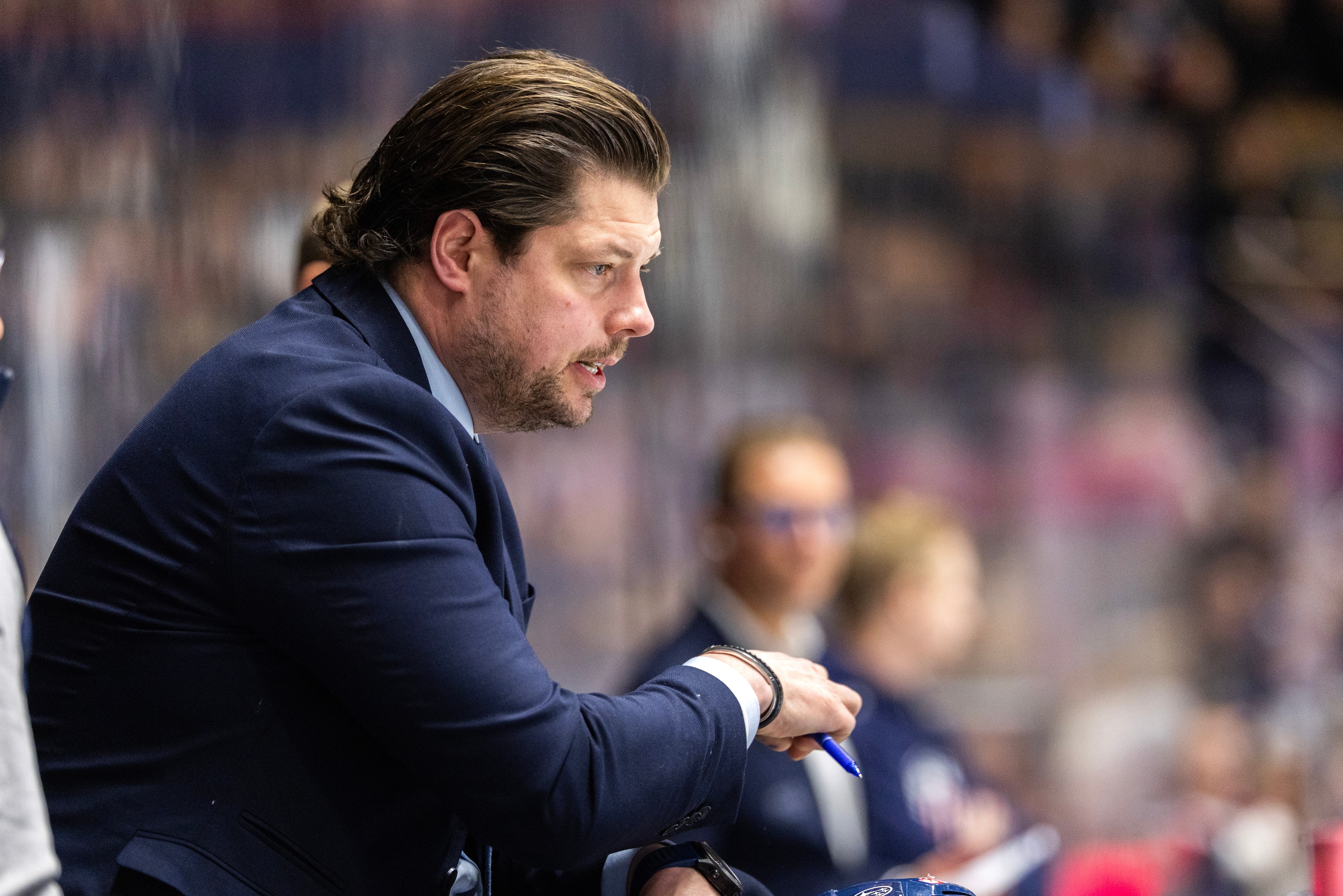 Man i mörk kostym sitter på bänken under en ishockeymatch och tittar fokuserat mot spelet.