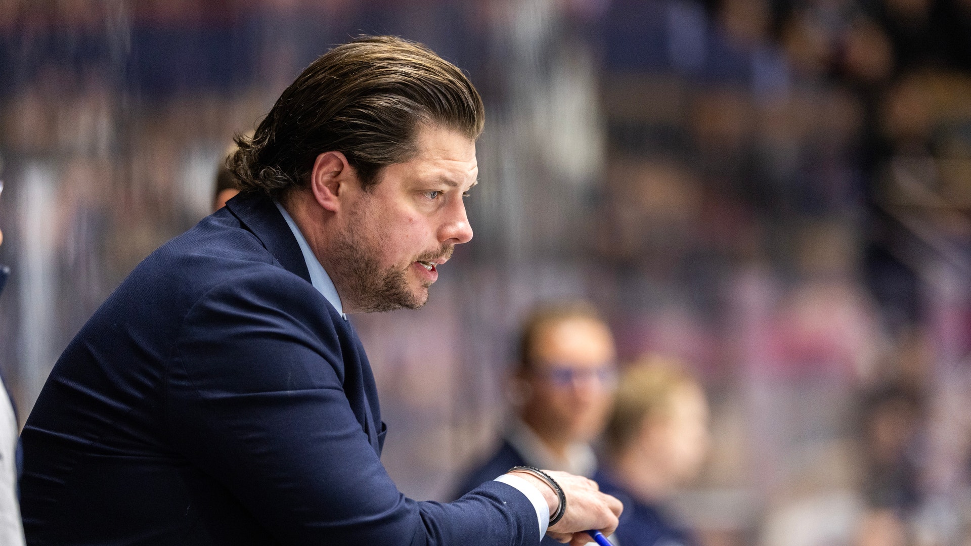 Man i mörk kostym sitter på bänken under en ishockeymatch och tittar fokuserat mot spelet.