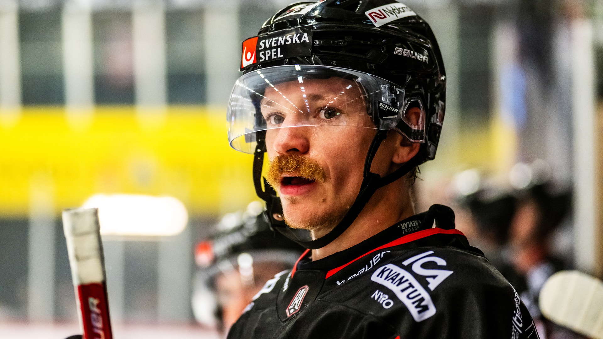 Porträtt av en ishockeyspelare i svart matchdräkt och hjälm med Svenska Spel-logga. Spelaren håller en ishockeyklubb och är fotograferad under en match.