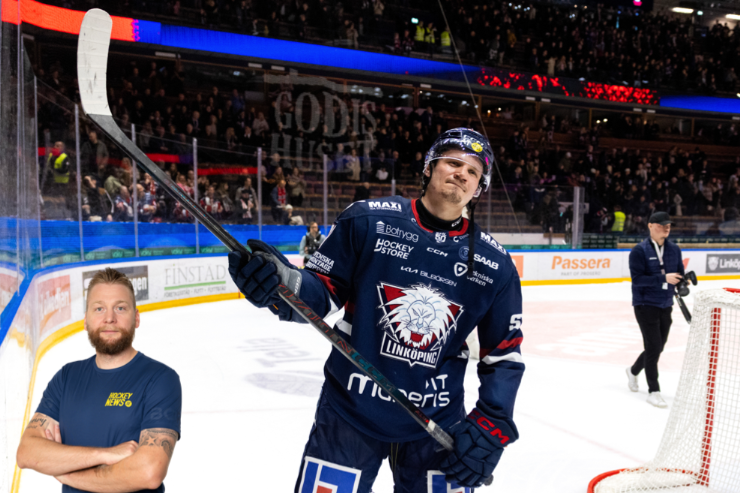 Ishockeyspelare i mörkblå tröja höjer sin klubba i luften på isen under en match. Publiken syns i bakgrunden tillsammans med bandor och arena-utrustning.