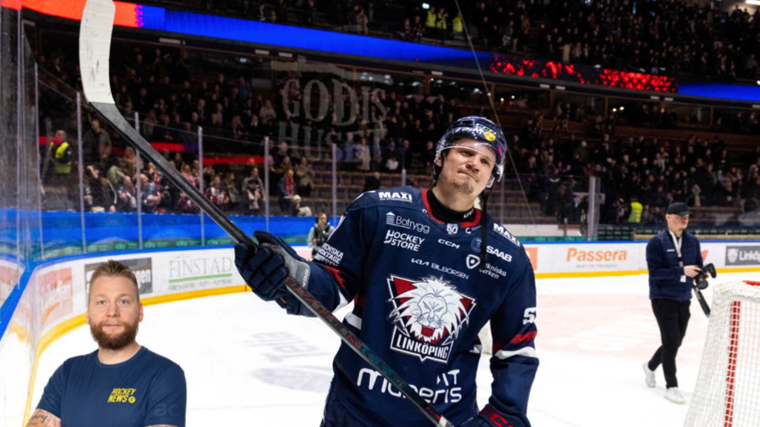 Ishockeyspelare i mörkblå tröja höjer sin klubba i luften på isen under en match. Publiken syns i bakgrunden tillsammans med bandor och arena-utrustning.