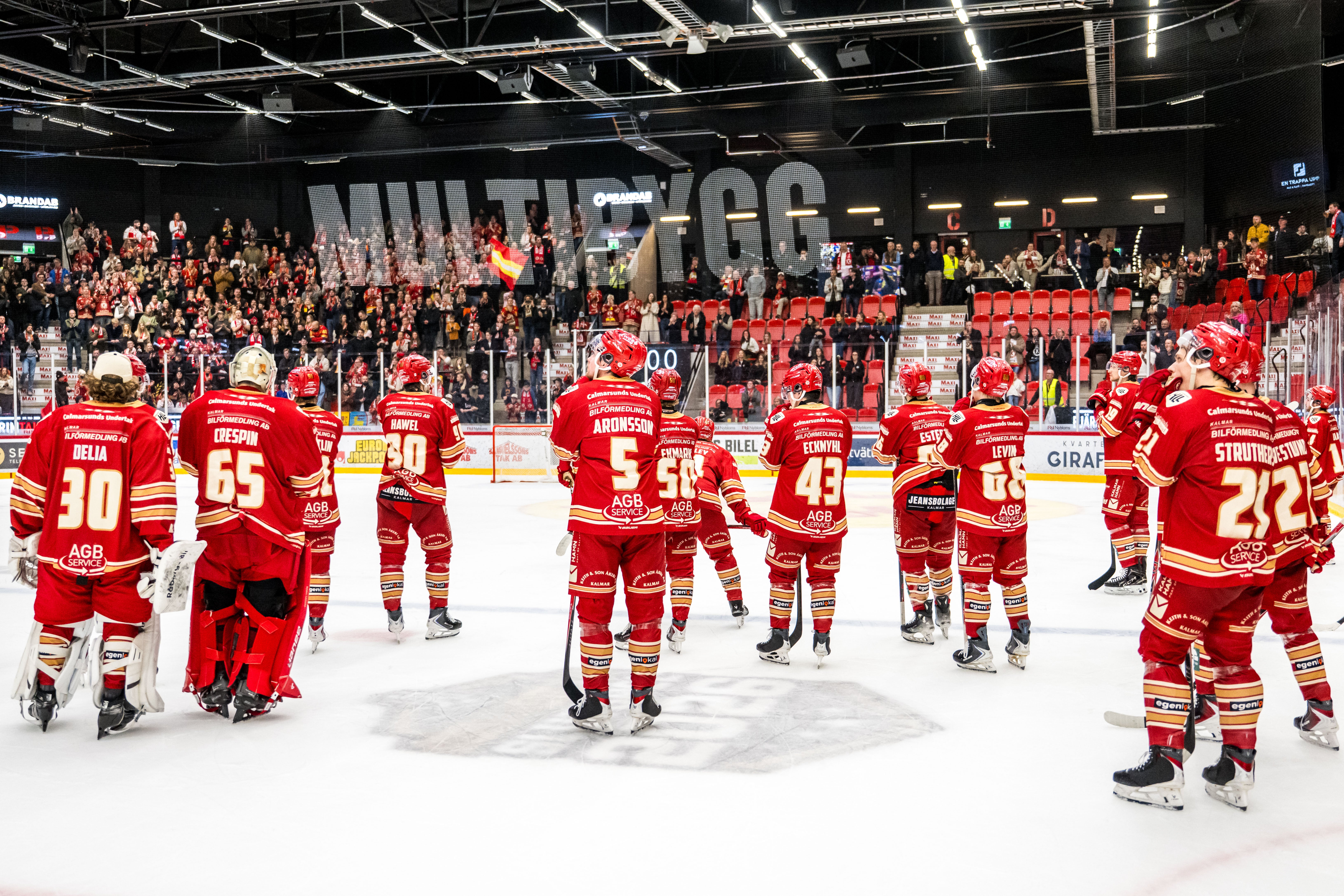 Ishockeylag i röda tröjor står på isen inför en match med fullsatt läktare i bakgrunden.