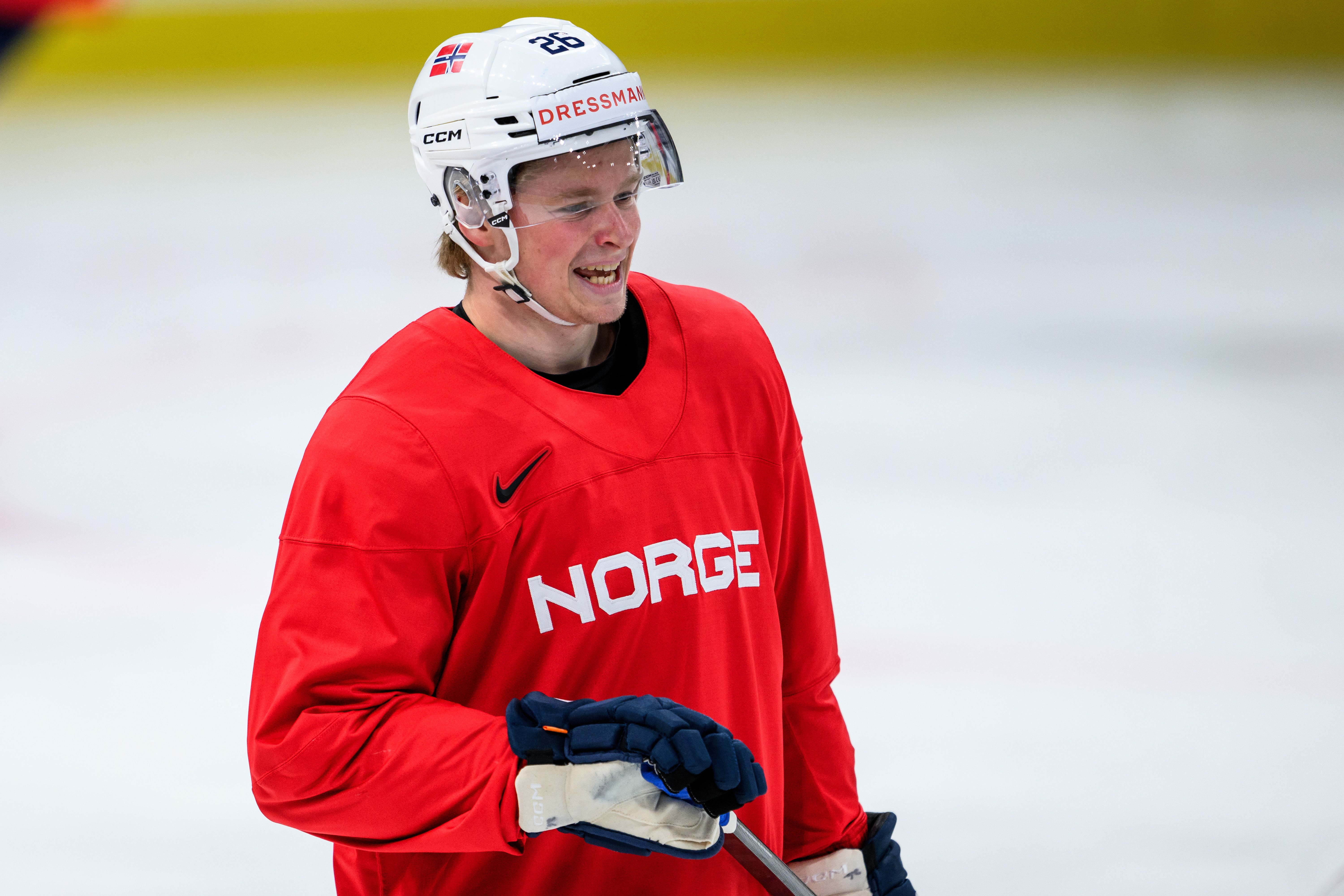 Ishockeyspelare i röd landslagströja med texten Norge, vit hjälm och blå handskar på isen. Ishockeyspelare i röda och gröna trikåer under en match