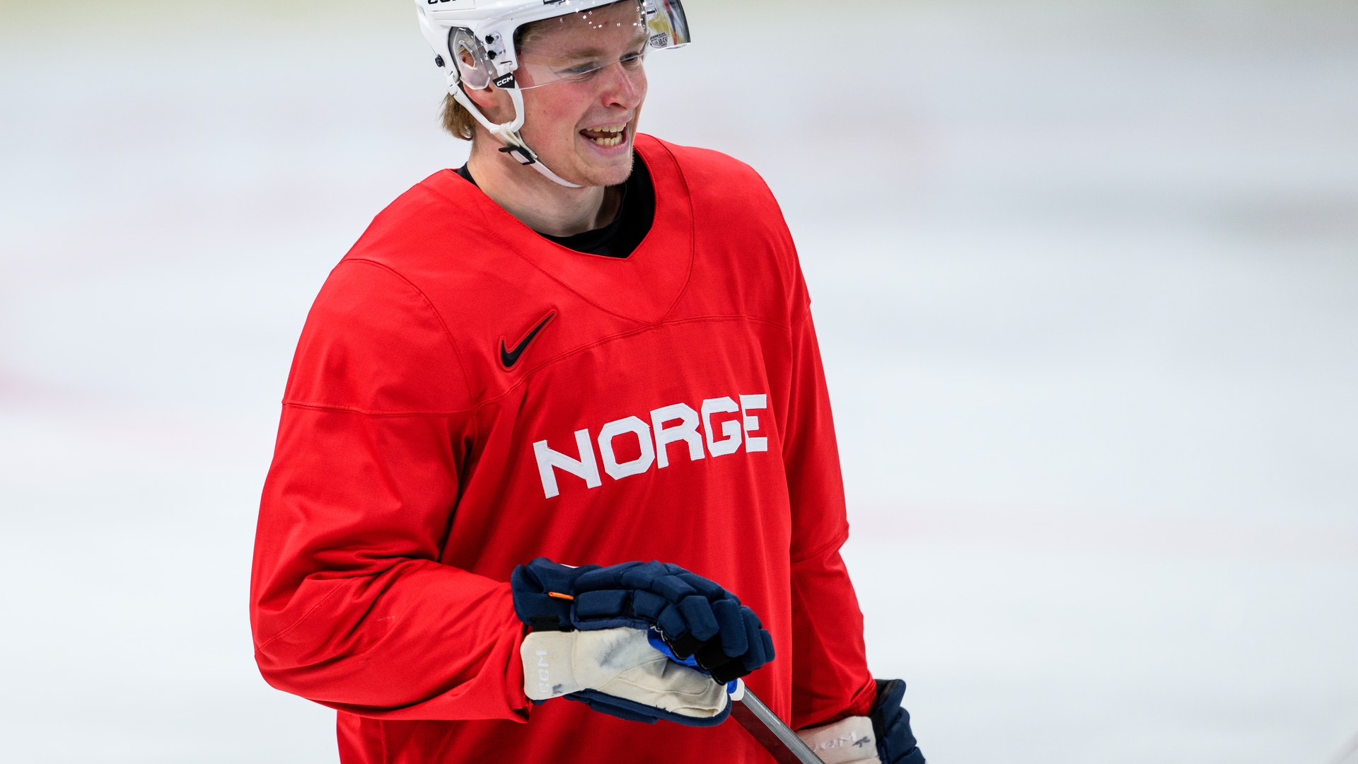 Ishockeyspelare i röd landslagströja med texten Norge, vit hjälm och blå handskar på isen. Ishockeyspelare i röda och gröna trikåer under en match