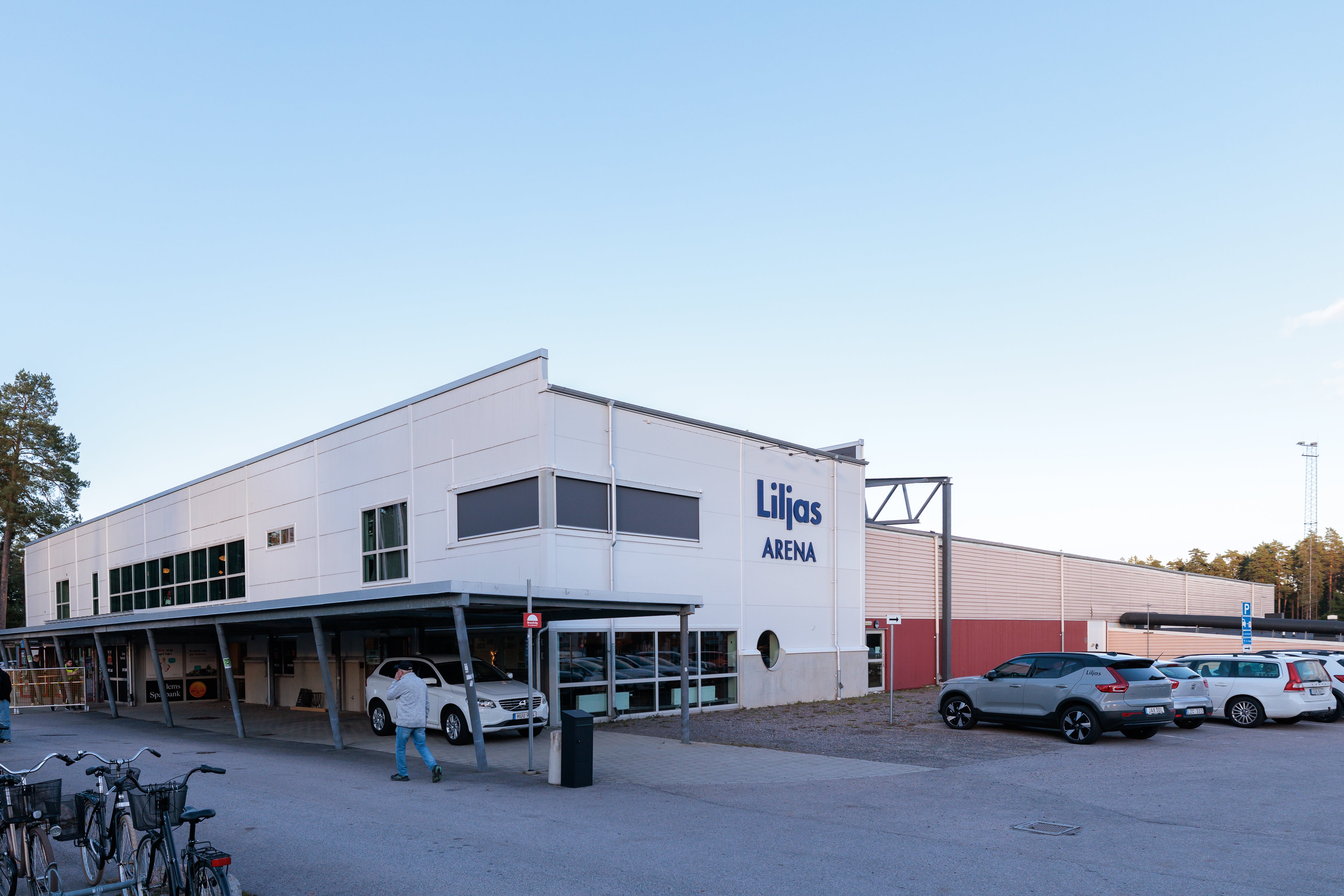 Exteriör av en modern vit byggnad med röda detaljer, märkt med Liljös Arena-logotyp. Byggnaden har stora fönster och är omgiven av en parkeringsplats med flera bilar parkerade framför.