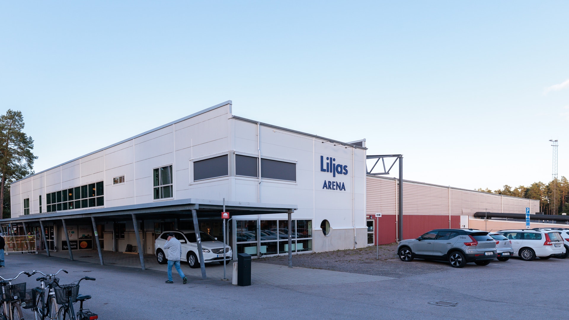 Exteriör av en modern vit byggnad med röda detaljer, märkt med Liljös Arena-logotyp. Byggnaden har stora fönster och är omgiven av en parkeringsplats med flera bilar parkerade framför.