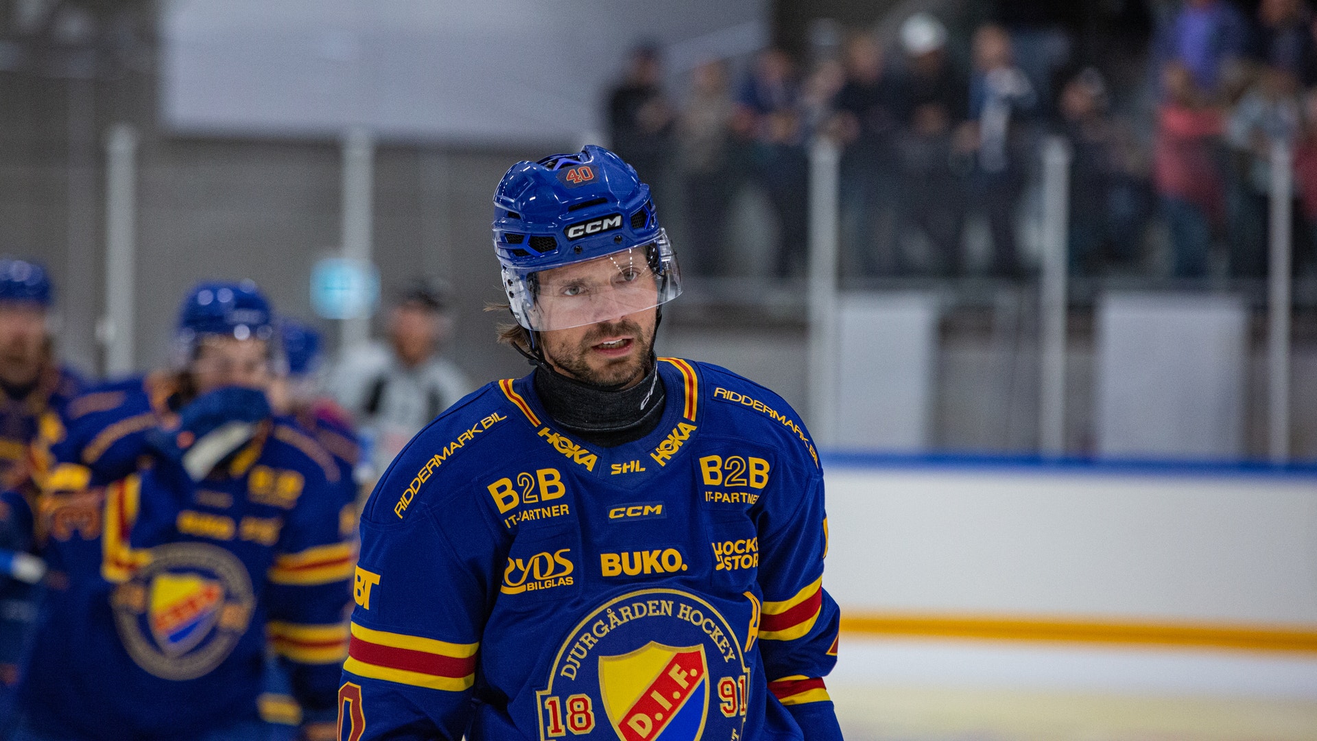Ishockeyspelare i blå och gul tröja står på isen under en match med publik i bakgrunden.