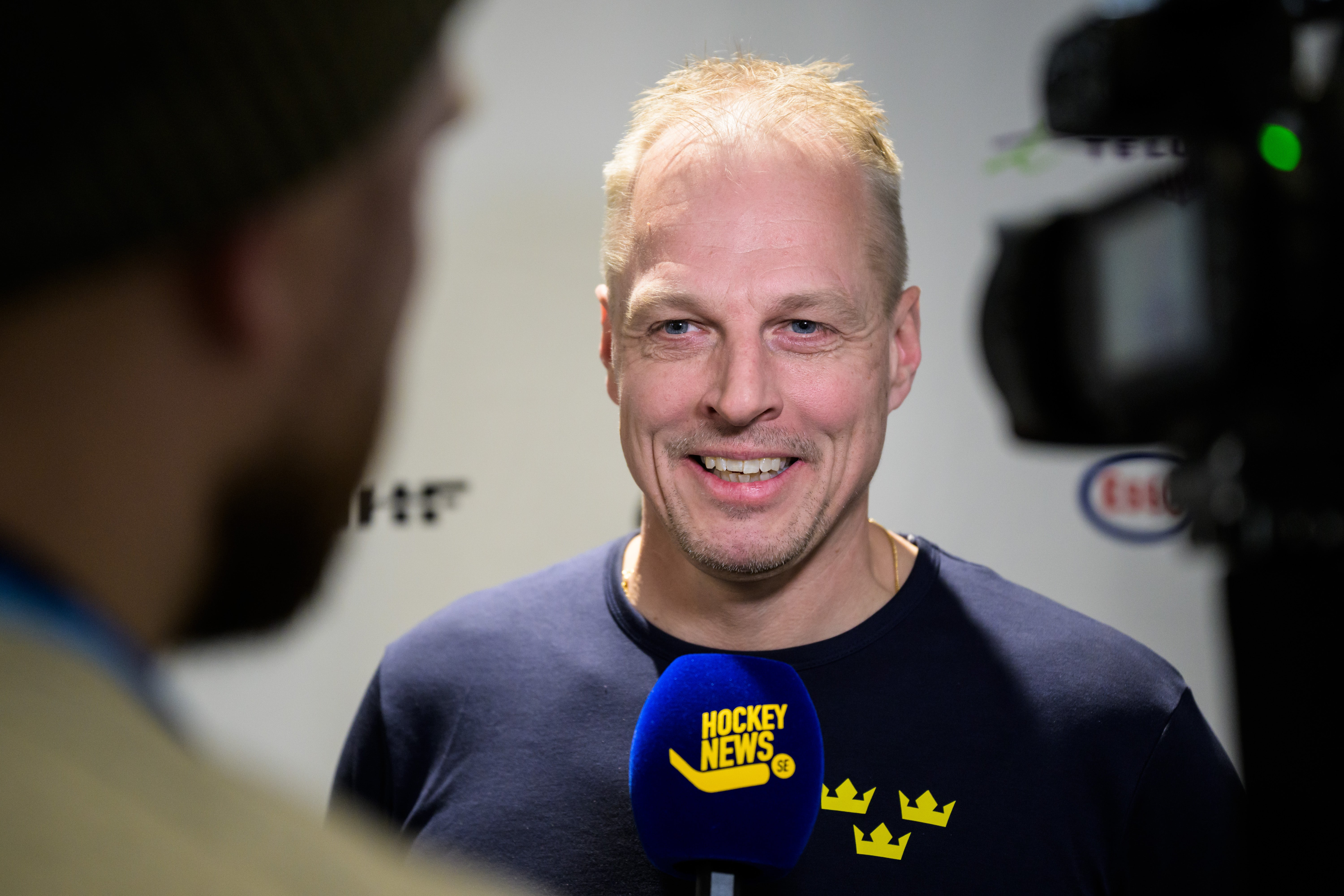 Man intervjuas framför kameror. Han bär en mörk tröja med Hockey News-logotyp och svenska flaggor.