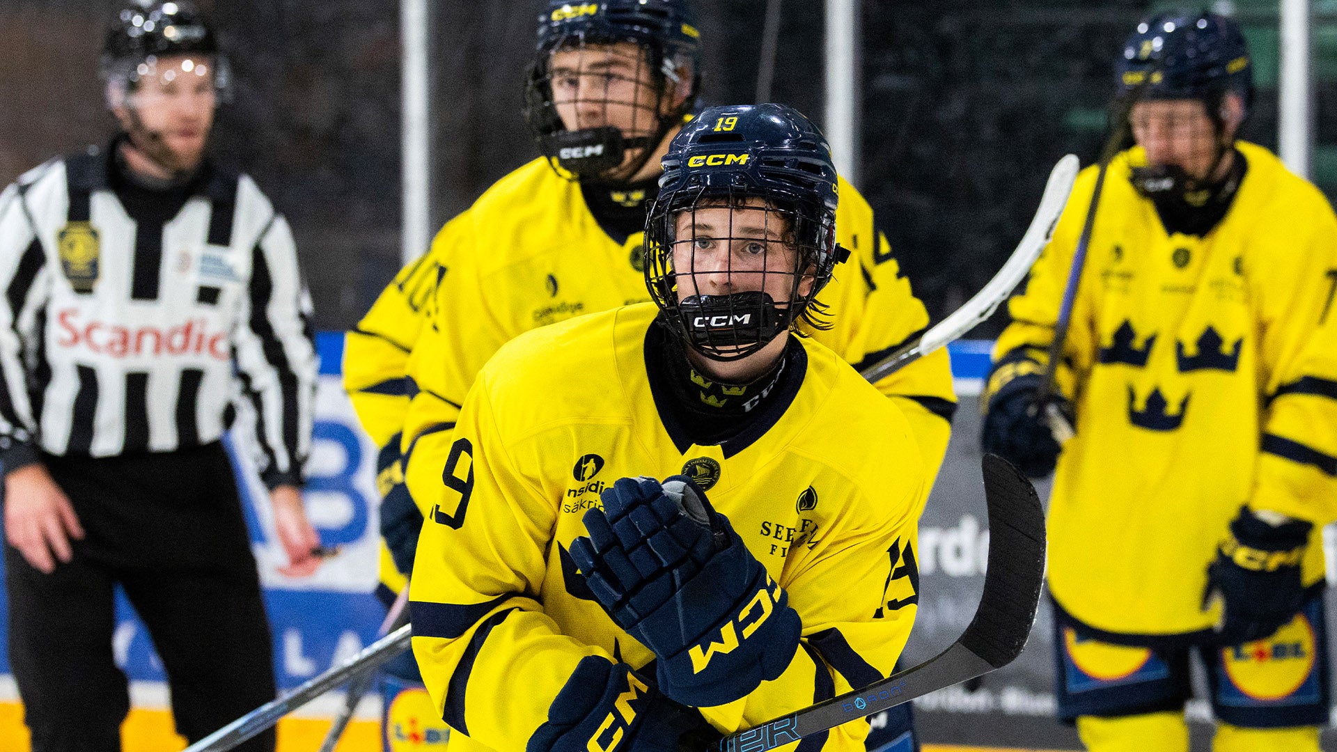 Ung ishockeyspelare i gul och blå svensk landslagsuniform spelar under en match. Spelaren är fokuserad på spelet med sin hockeyklubb i händerna. En domare i svartvit randig uniform och andra spelare syns i bakgrunden.