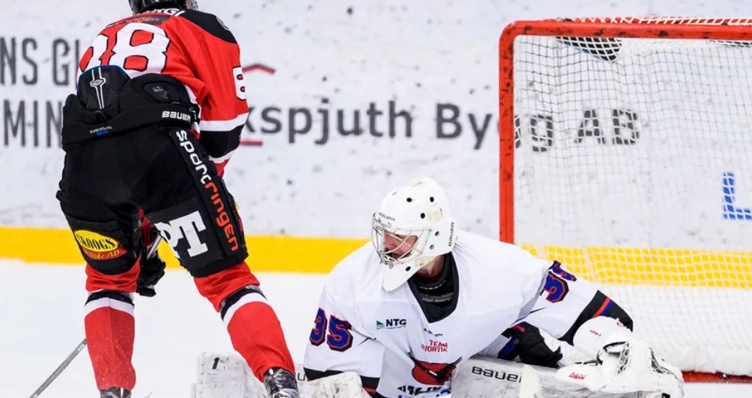 En målvakt i vit matchdräkt håller vakt vid målet under en ishockeymatch, medan en spelare i röd tröja är nära.
