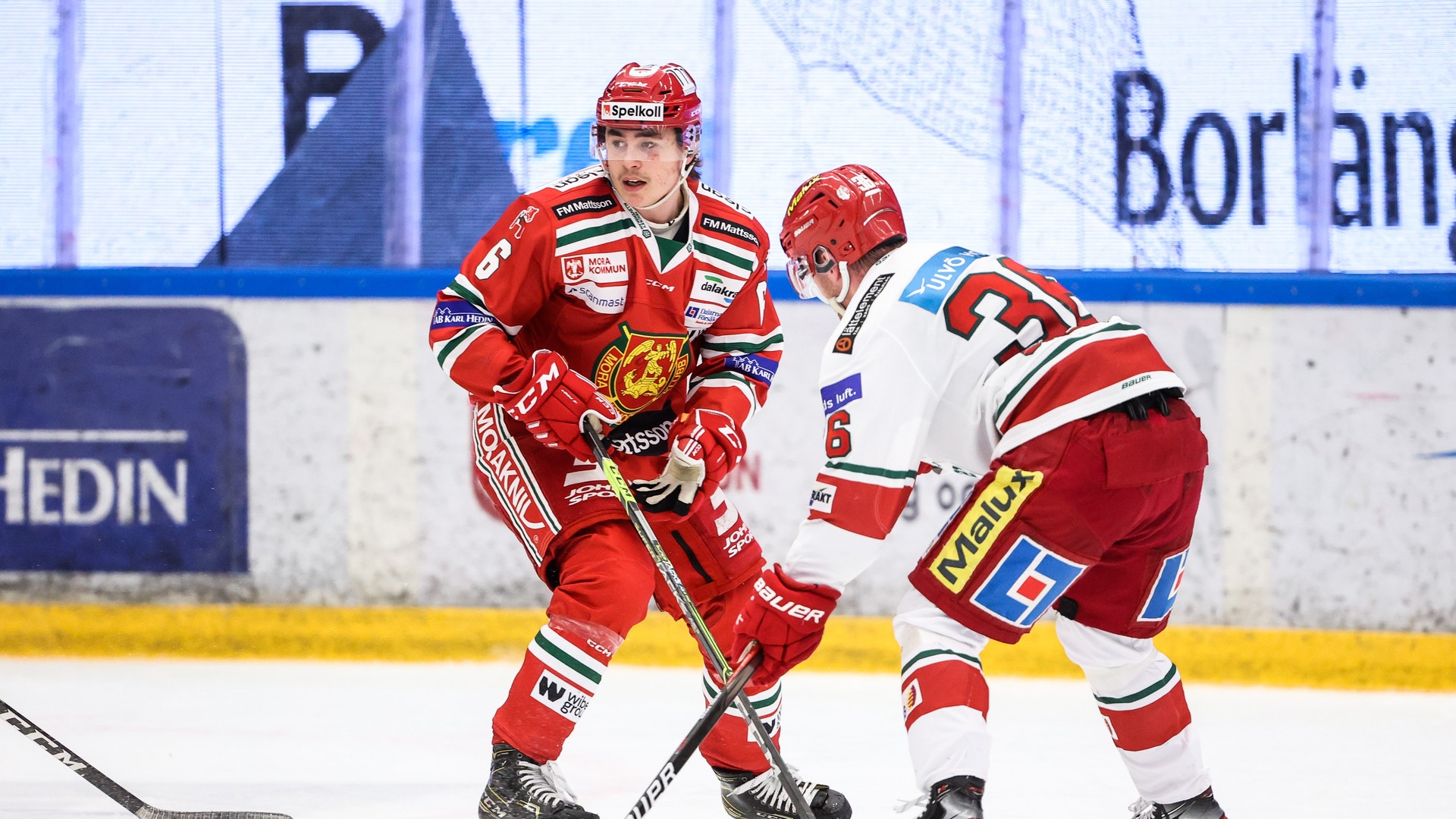 Två ishockeyspelare i aktion på isen under en match. Spelaren i röd tröja och spelaren i vit tröja med röda detaljer kämpar om pucken med sina klubbor. Publiken och bandorna syns i bakgrunden.