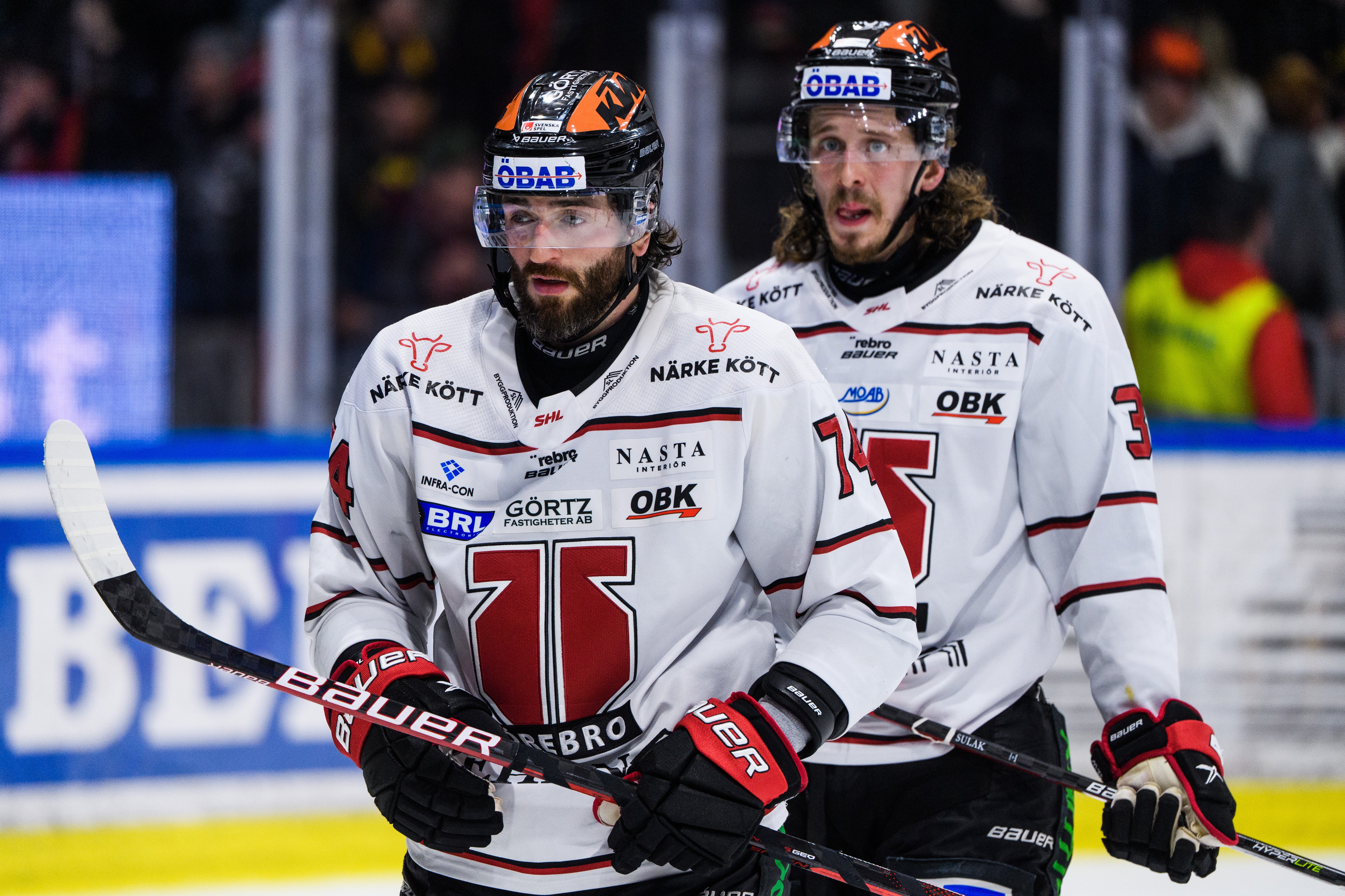 Två ishockeyspelare i vita och röda trikåer står tillsammans på isen under en match. De bär hjälmar och handskar och ser fokuserade ut.