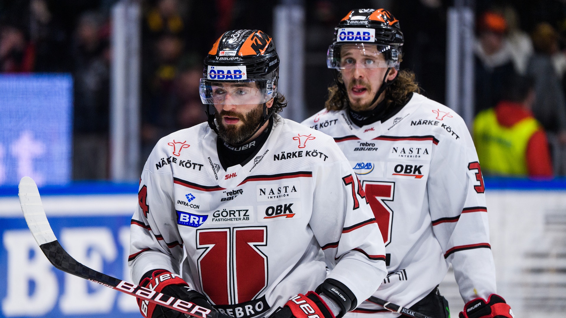 Två ishockeyspelare i vita och röda trikåer står tillsammans på isen under en match. De bär hjälmar och handskar och ser fokuserade ut.
