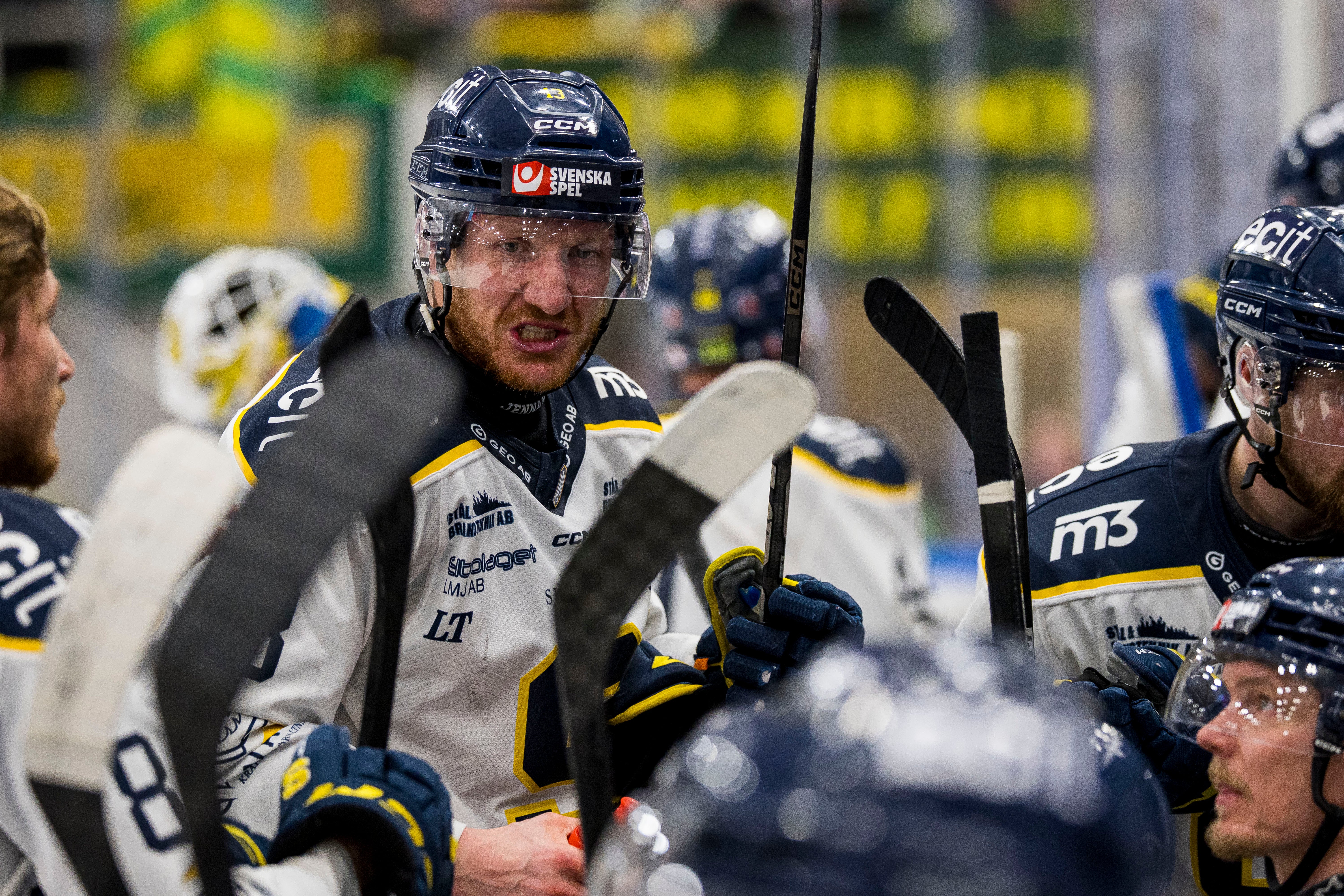 Ishockeymatch där en spelare i svart och vit tröja med gula detaljer står på bänken tillsammans med sina lagkamrater och håller sin hockeyklubbe.