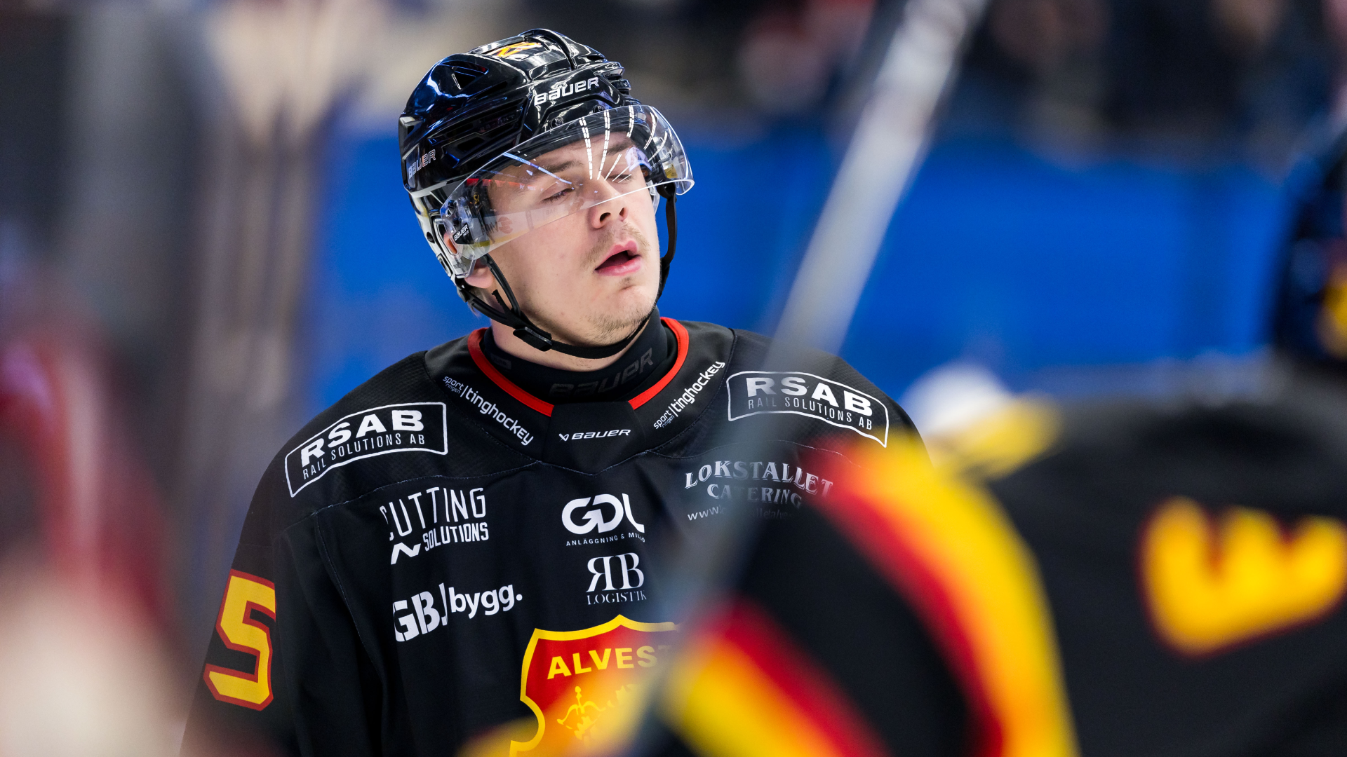 En ishockeyspelare i svart tröja och hjälm med visir blickar nedåt under en match.