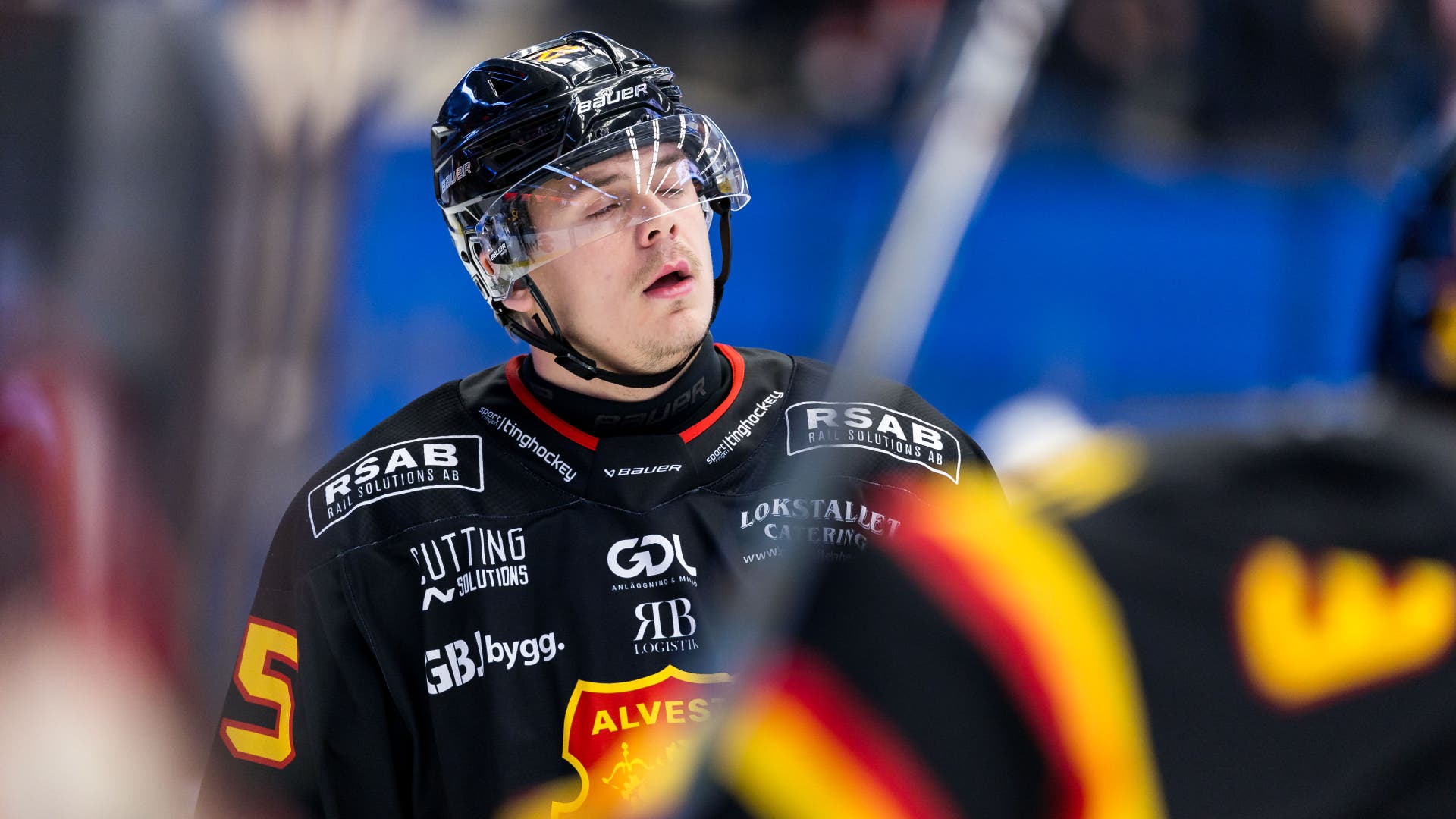 En ishockeyspelare i svart tröja och hjälm med visir blickar nedåt under en match.