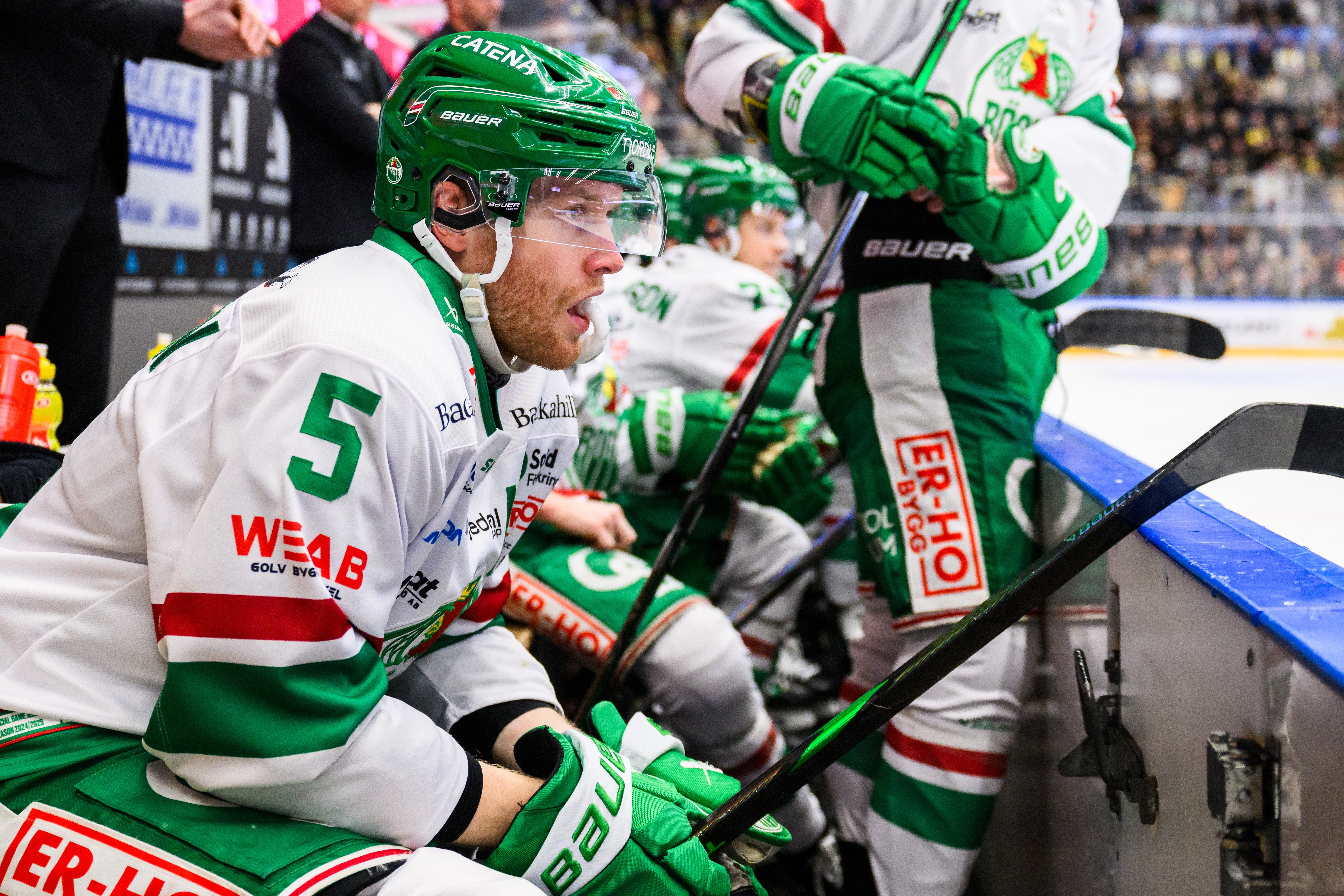 Ishockeyspelare i grön och vit matchdräkt sitter på spelarnas bänk under en match. Ishockeyspelare i vit och grön matchdräkt med hjälm under en match i en stor arena med publik i bakgrunden.