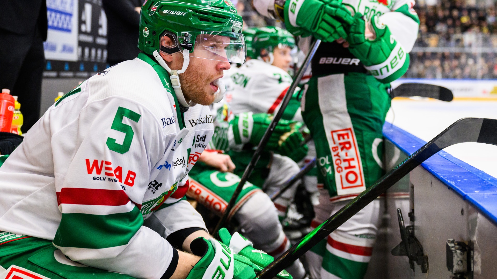 Ishockeyspelare i grön och vit matchdräkt sitter på spelarnas bänk under en match. Ishockeyspelare i vit och grön matchdräkt med hjälm under en match i en stor arena med publik i bakgrunden.
