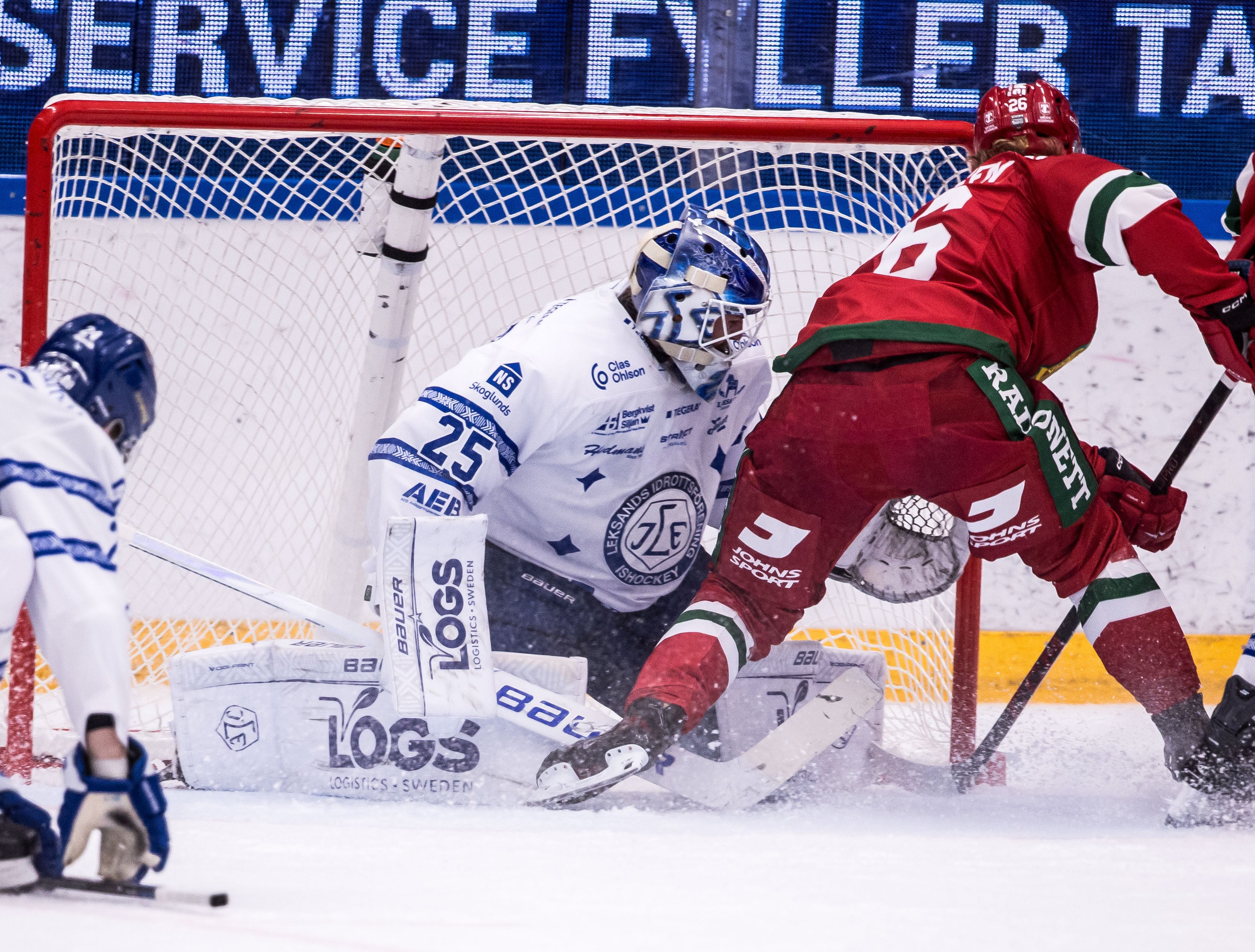 En målvakt räddar ett skott framför mål under en ishockeymatch, med en anfallande spelare i rött nära kassen.
