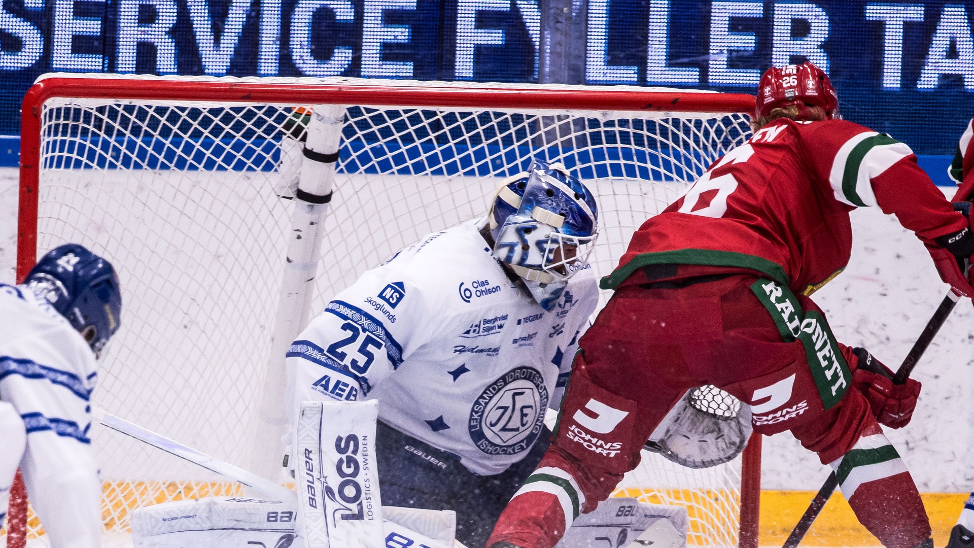 En målvakt räddar ett skott framför mål under en ishockeymatch, med en anfallande spelare i rött nära kassen.