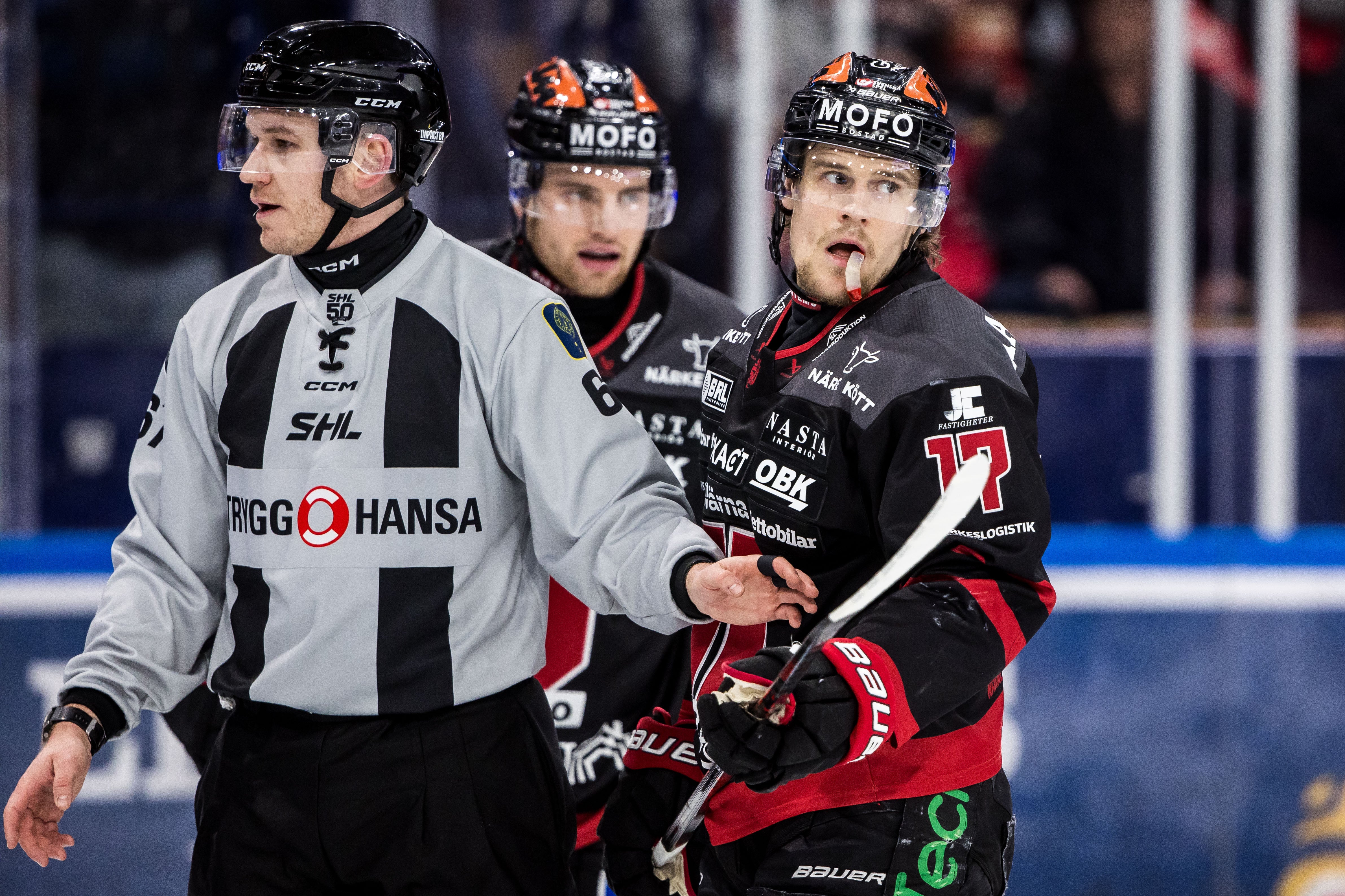 Tre ishockeyspelare under en match. En spelare i vit och svart Hansa-tröja står på vänster sida, medan två spelare i svart och röd tröja är positionerade på höger sida av bilden.