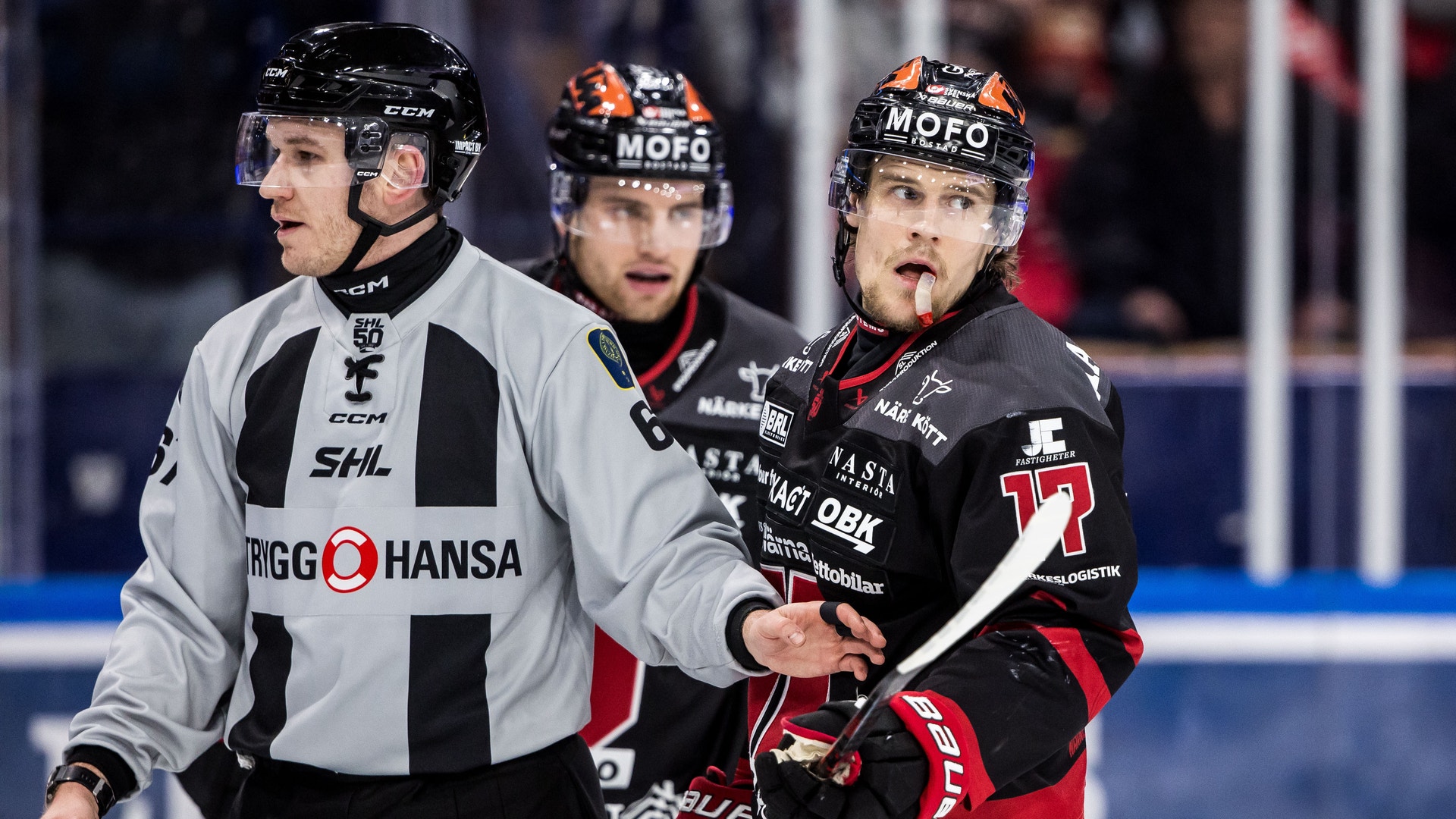 Tre ishockeyspelare under en match. En spelare i vit och svart Hansa-tröja står på vänster sida, medan två spelare i svart och röd tröja är positionerade på höger sida av bilden.