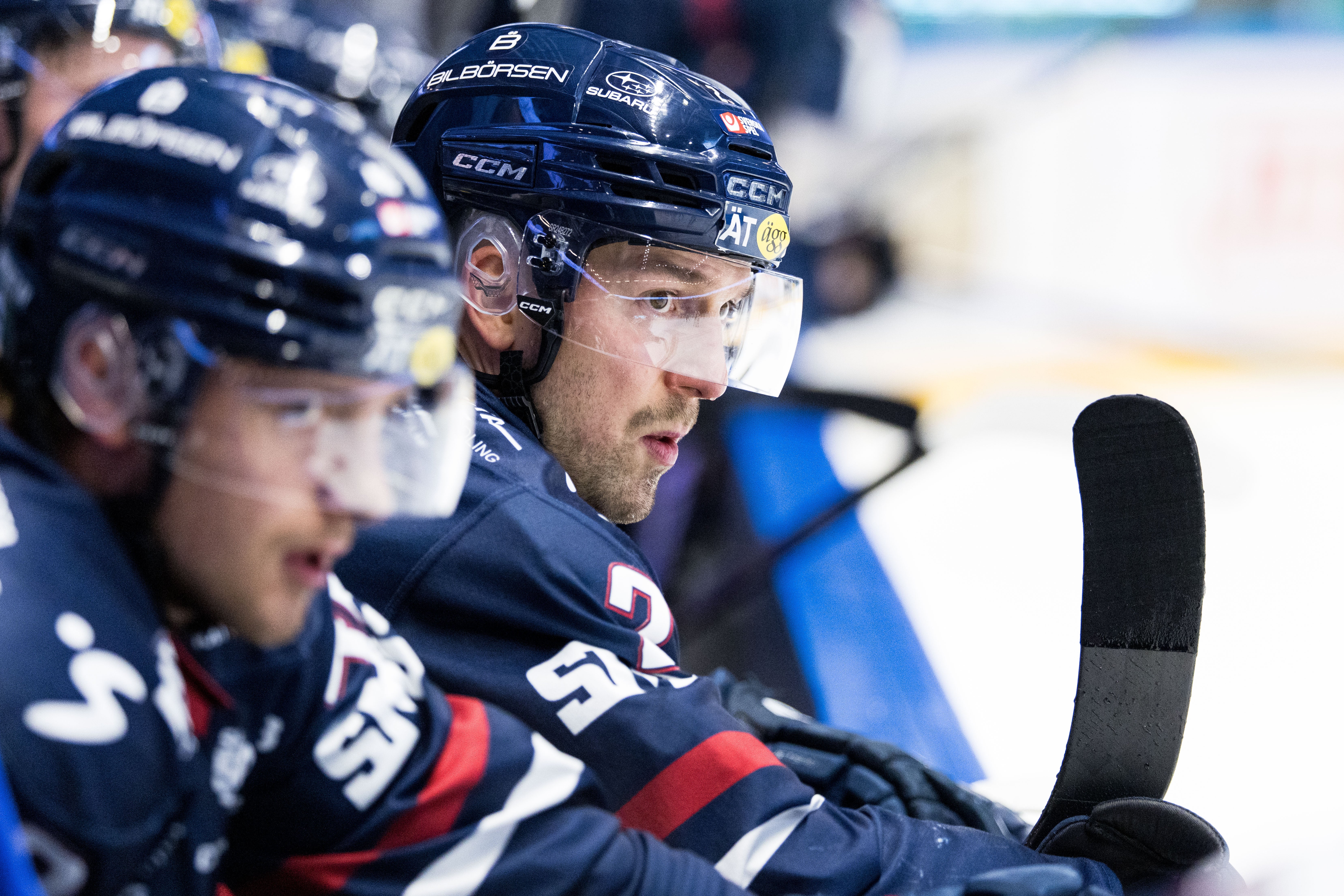 Ishockeyspelare i mörkblå lag sitter på bänken under en match. Spelaren i förgrunden bär hjälm och ser fokuserad ut, med en hockeyklubba synlig i bilden.