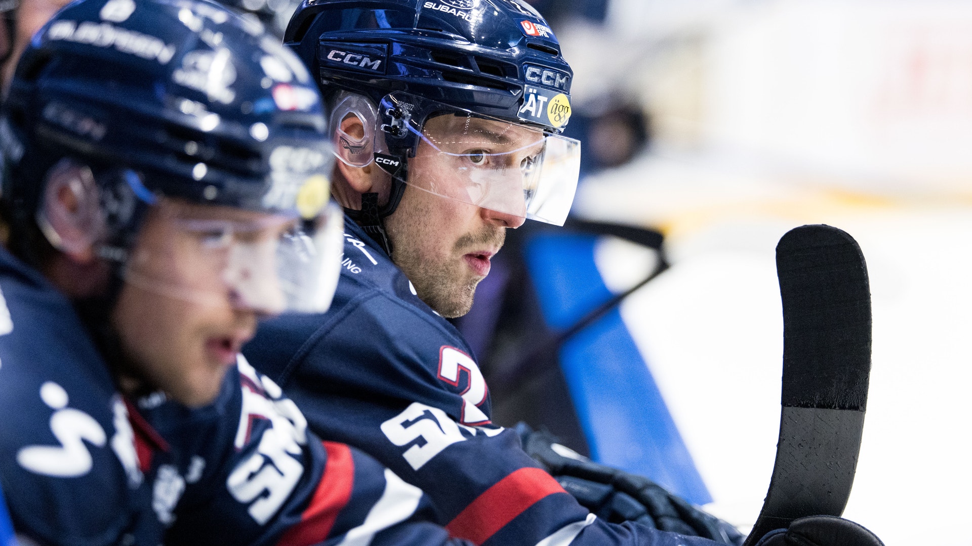 Ishockeyspelare i mörkblå lag sitter på bänken under en match. Spelaren i förgrunden bär hjälm och ser fokuserad ut, med en hockeyklubba synlig i bilden.